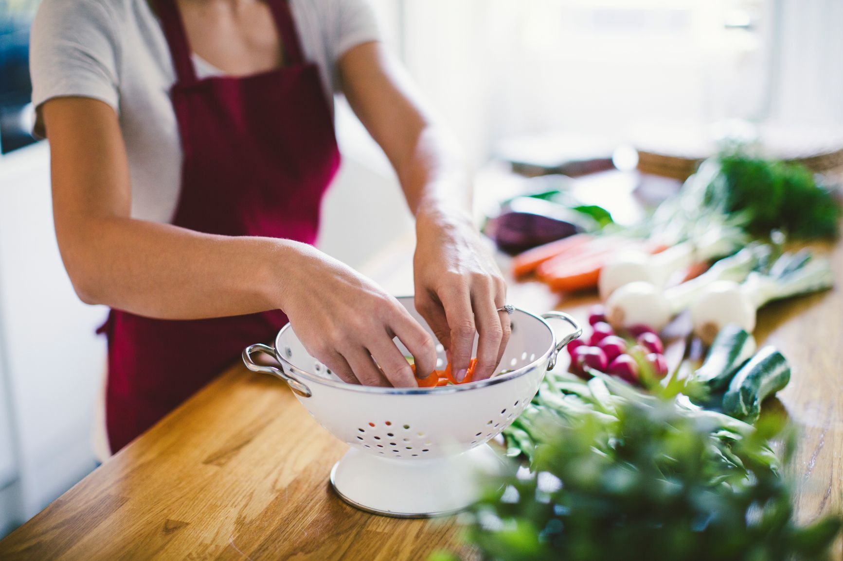 A person wearing a maroon apron washes fresh vegetables in a white colander on a wooden kitchen counter.