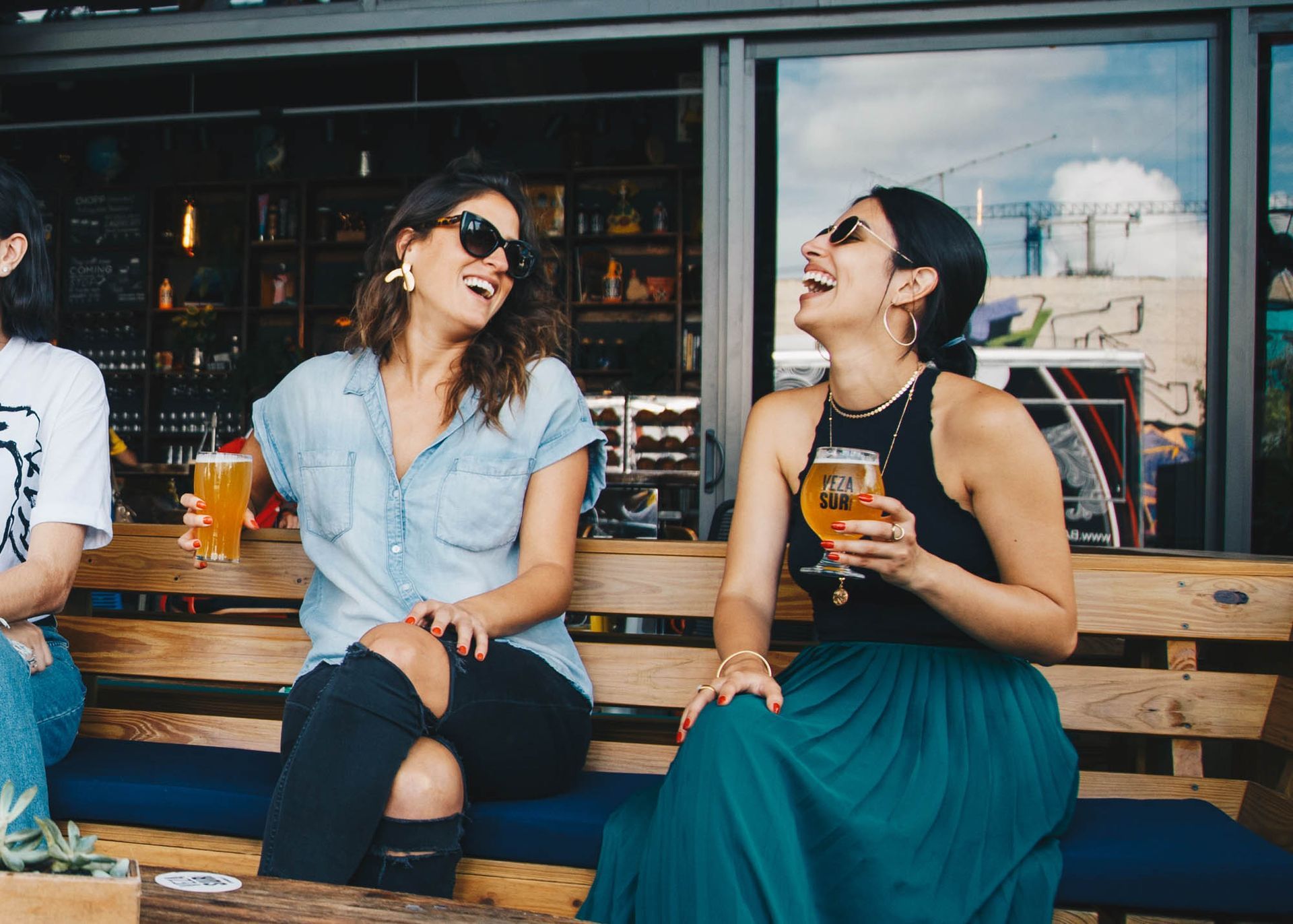 Three people laugh while sitting on a wooden bench outdoors, holding drinks in a social setting.