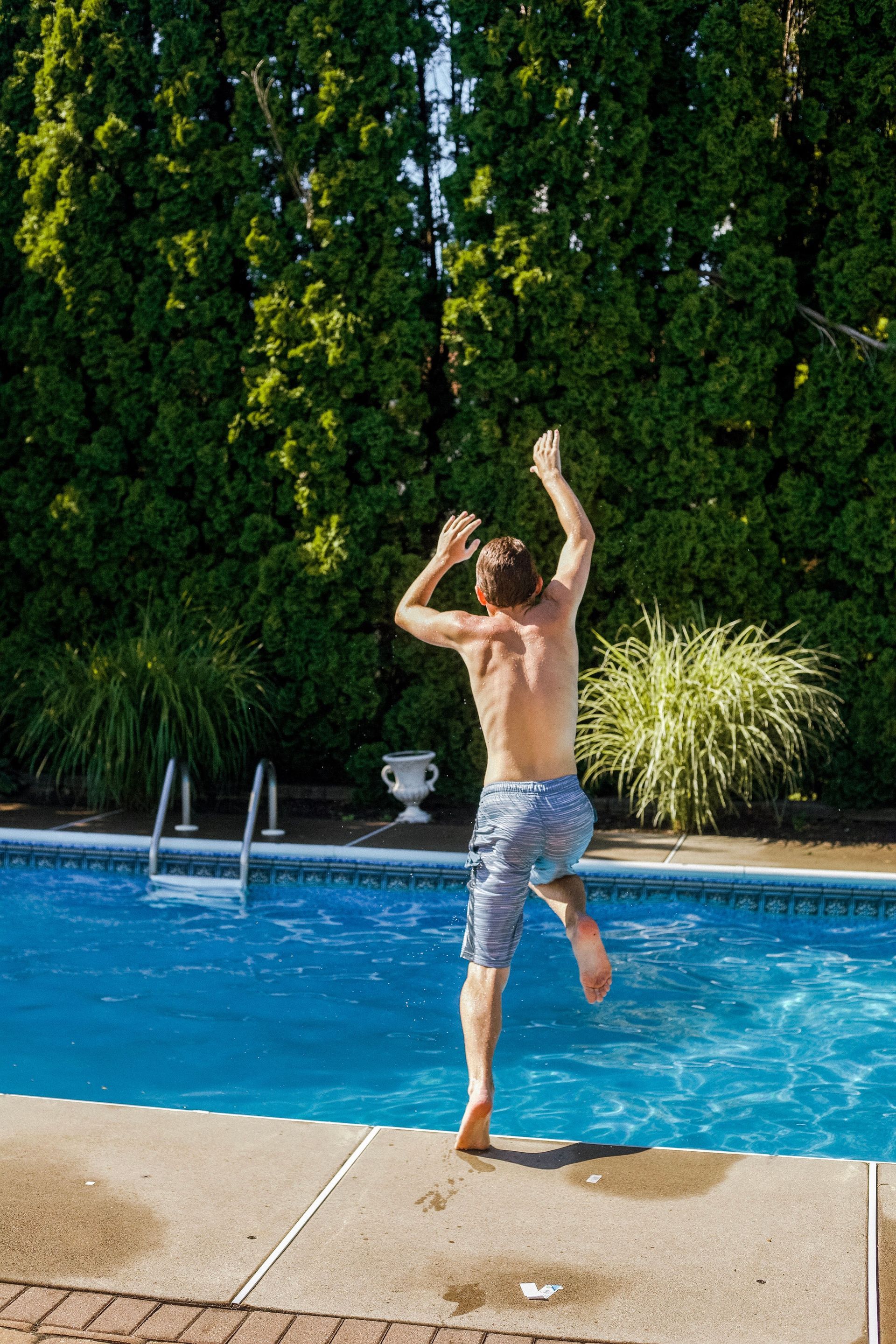 Man jumping into a backyard pool on a sunny day, with blue water and tall greenery behind him