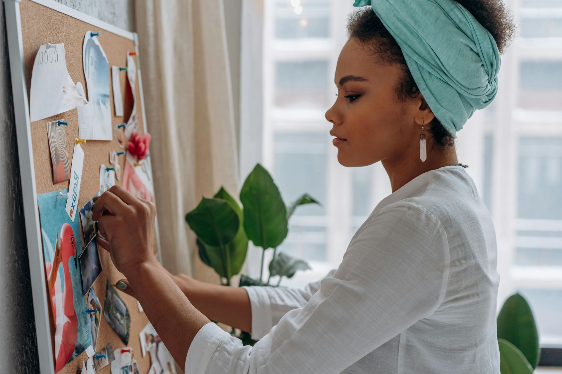 A person wearing a light blue headwrap and white shirt pins a paper to a cork inspiration board in a bright room.