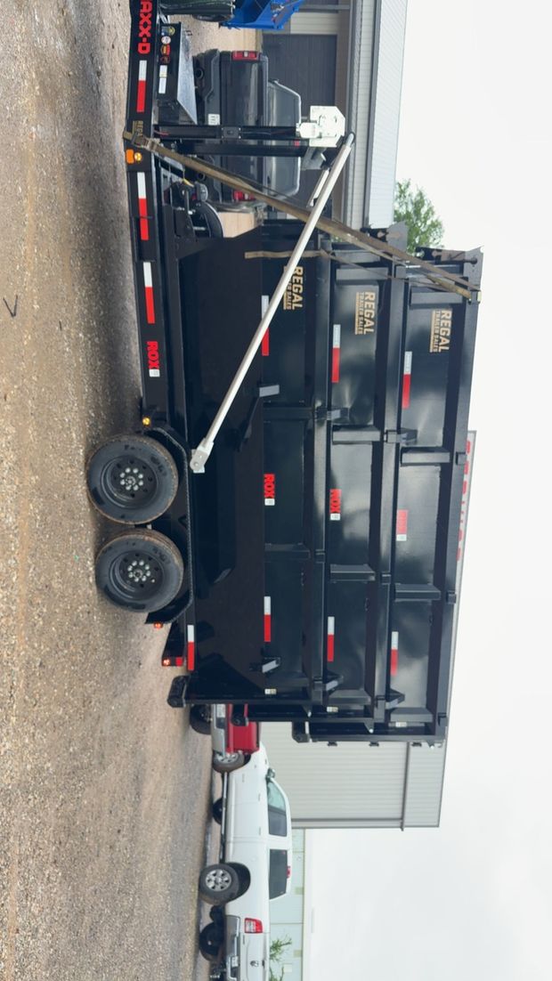 Black dump trailer with red accents parked on a gravel lot beside a white truck.