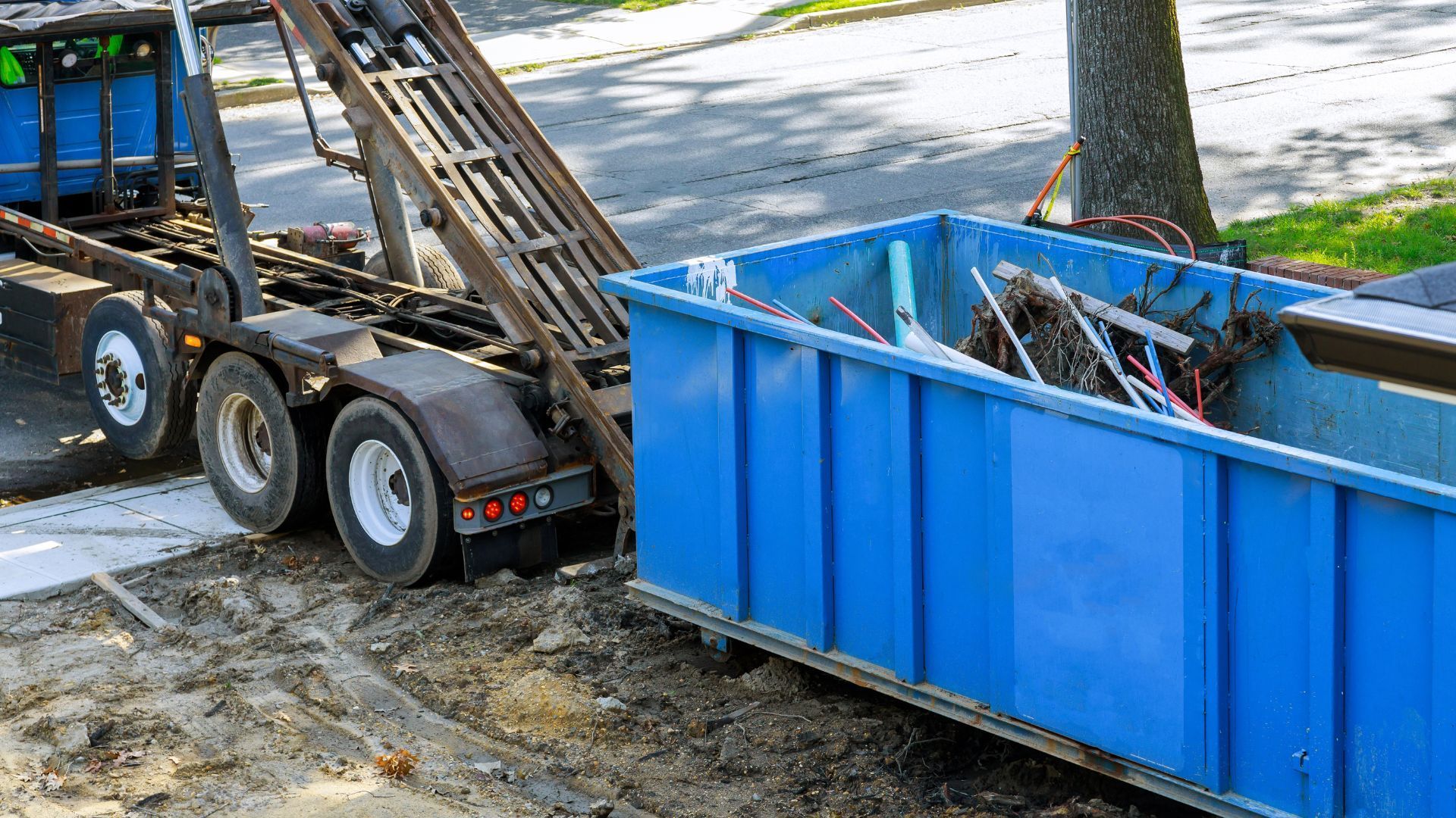 Blue dumpster filled with debris beside a trailer on a dirt roadside driveway.