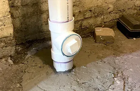 White PVC pipe with round cleanout cap in a muddy basement corner against a rough concrete wall