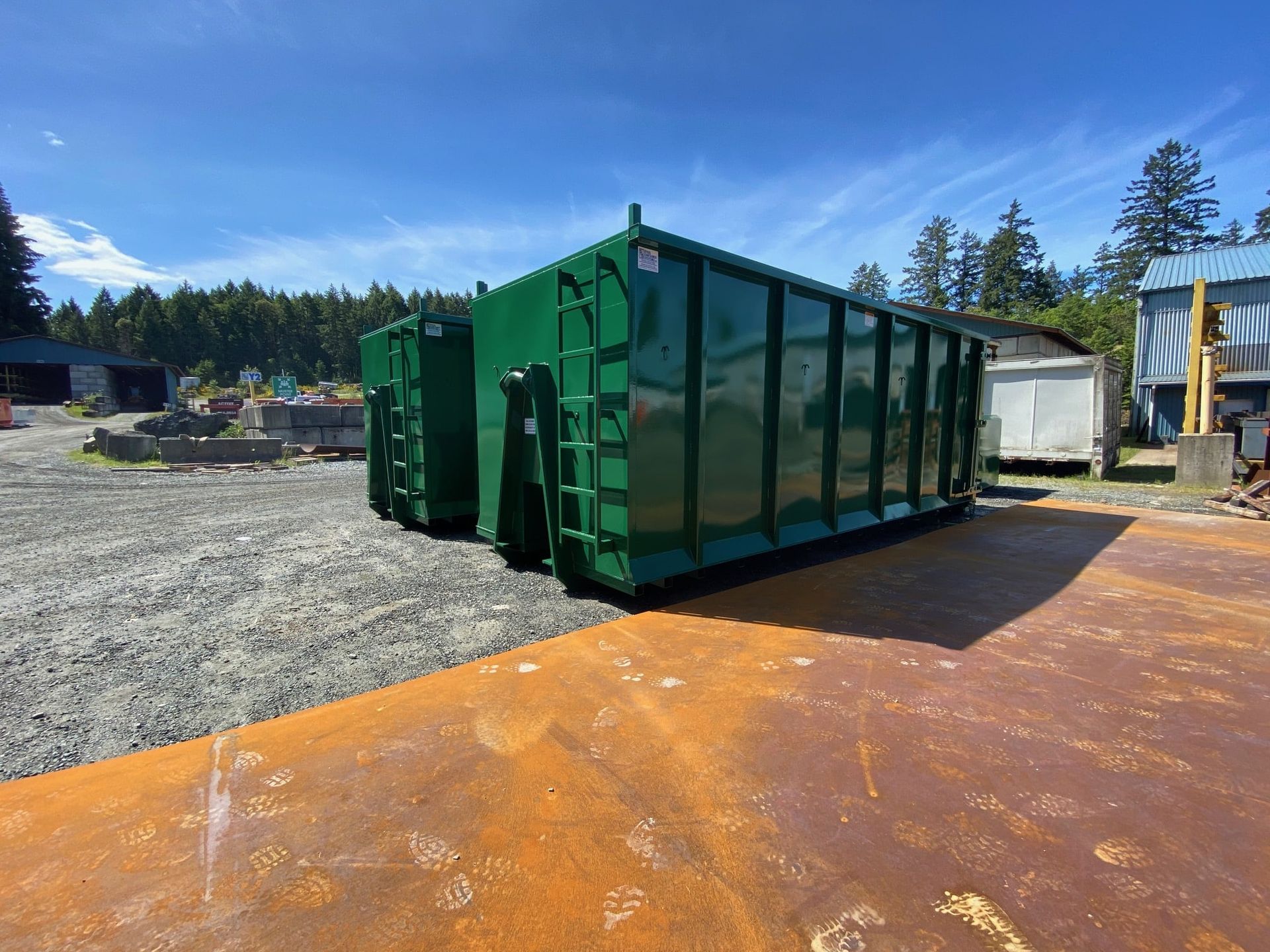 Large green industrial dumpster on gravel lot with rust-colored ground and trees in the background