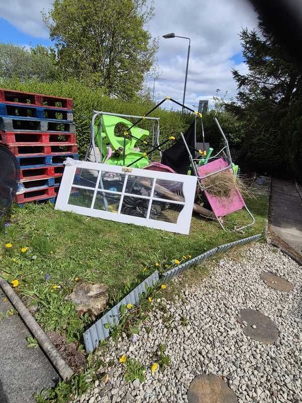 Colorful pallet art installation on a grassy roadside, with framed panels and chairs amid trees and gravel.
