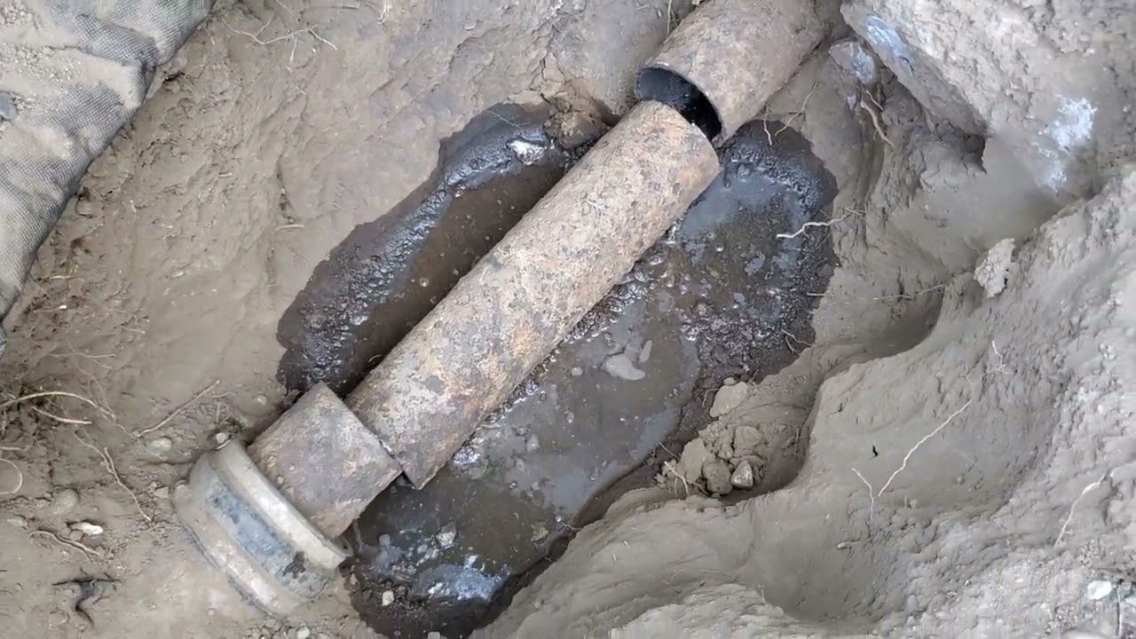 Buried water pipe junction in a muddy excavation pit, with dark wet soil around it