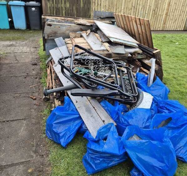 Pile of scrap wood, metal, and blue bags stacked beside a fence outdoors.