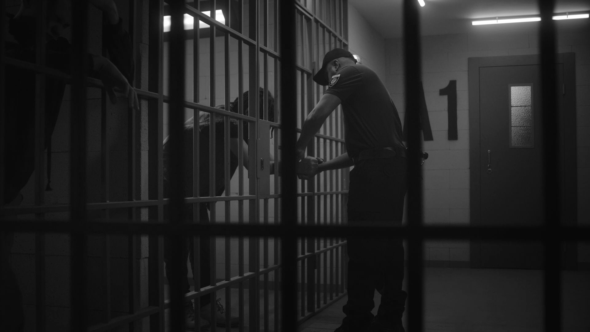 Person leaning over prison cell bars toward another inmate in a dim jail corridor.