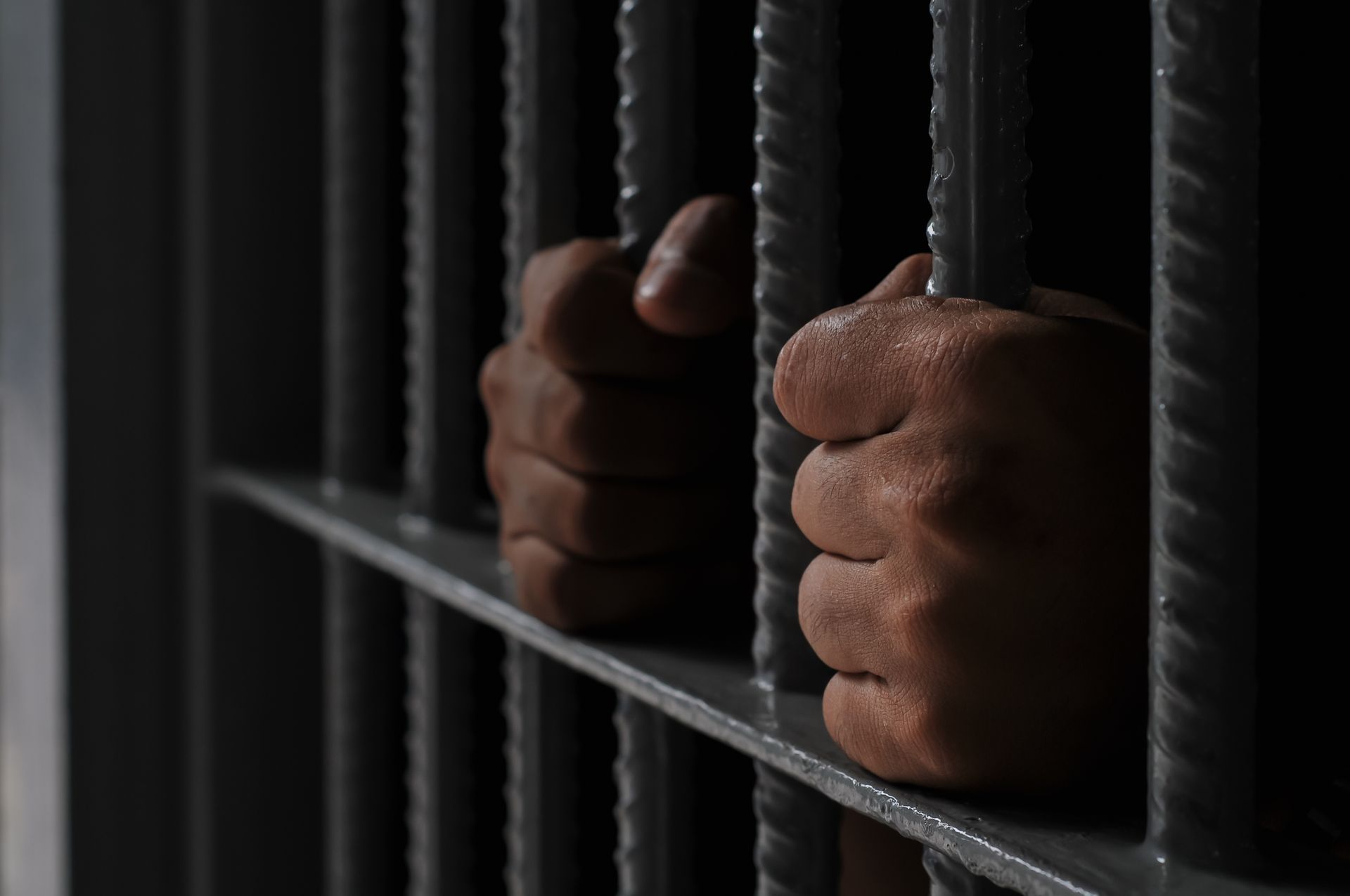 Hands gripping prison bars in a dark jail cell.
