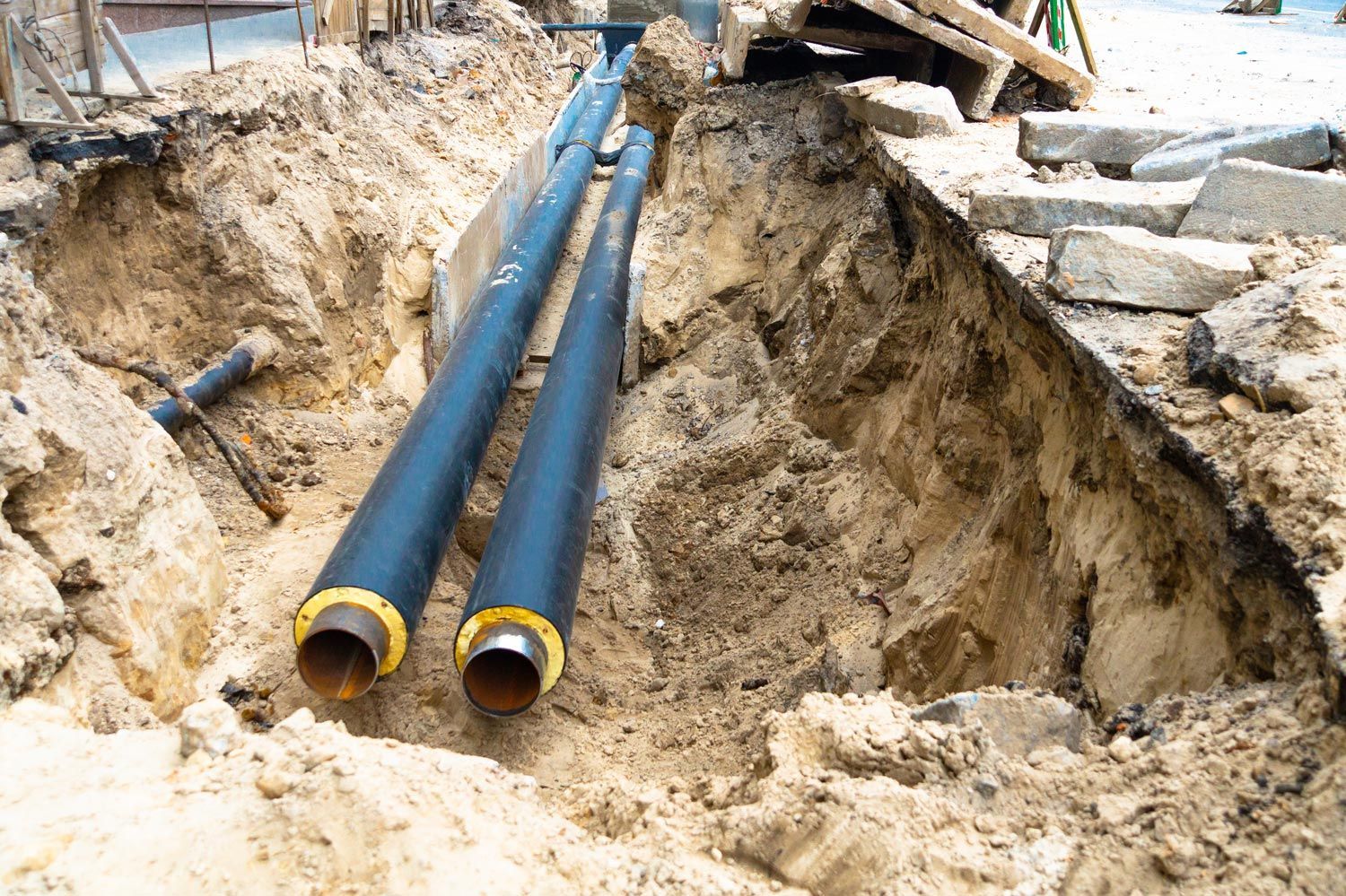 Two black insulated pipes running through a narrow, open dirt trench at a construction site.