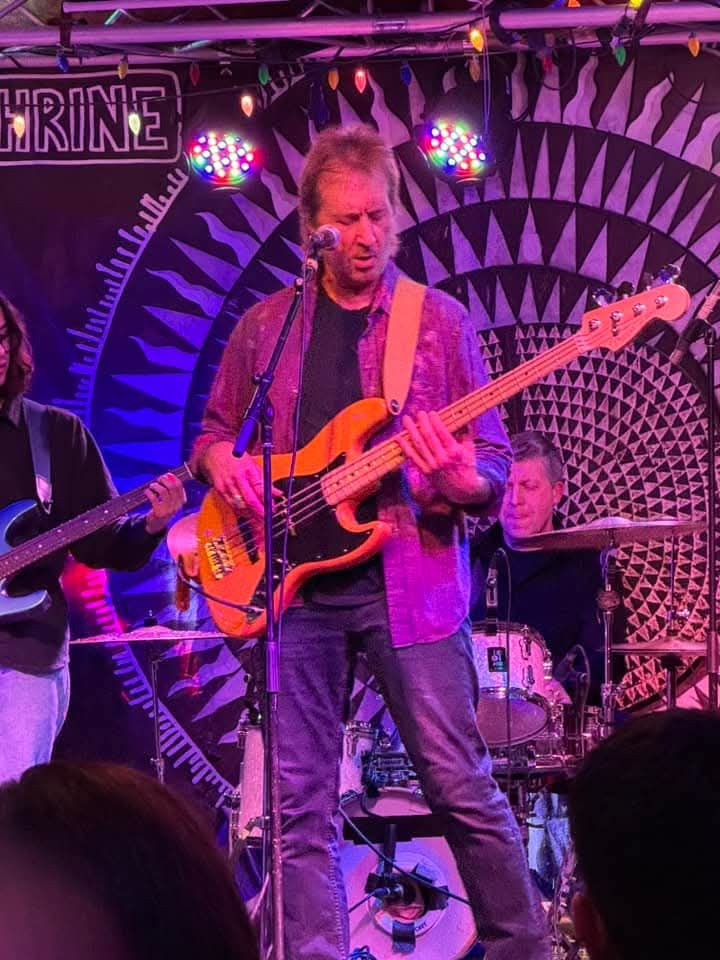 Musician playing bass guitar on a purple-lit stage with a drummer in the background