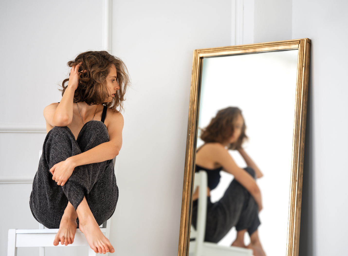 Woman sitting on a stool beside a gold-framed mirror, looking at her reflection in a bright room