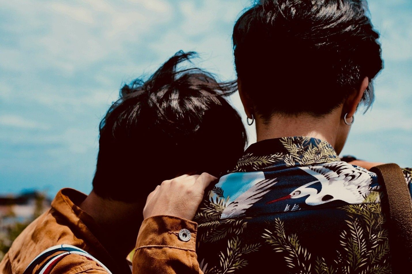 Two people standing close together outdoors, seen from behind under a blue sky.