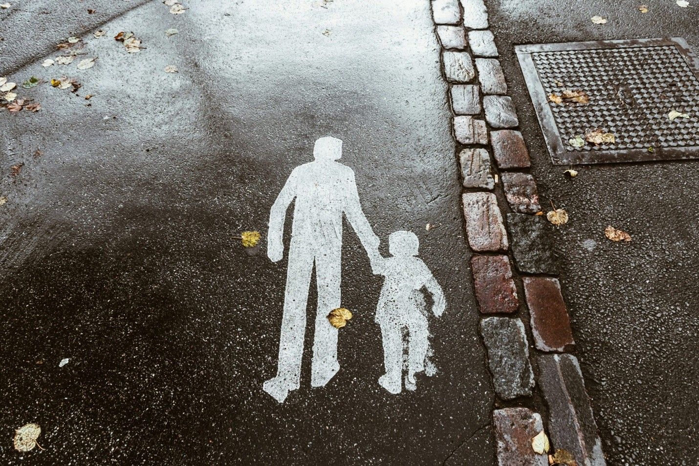 White silhouette of an adult holding a child's hand on a sidewalk near a curb and manhole cover