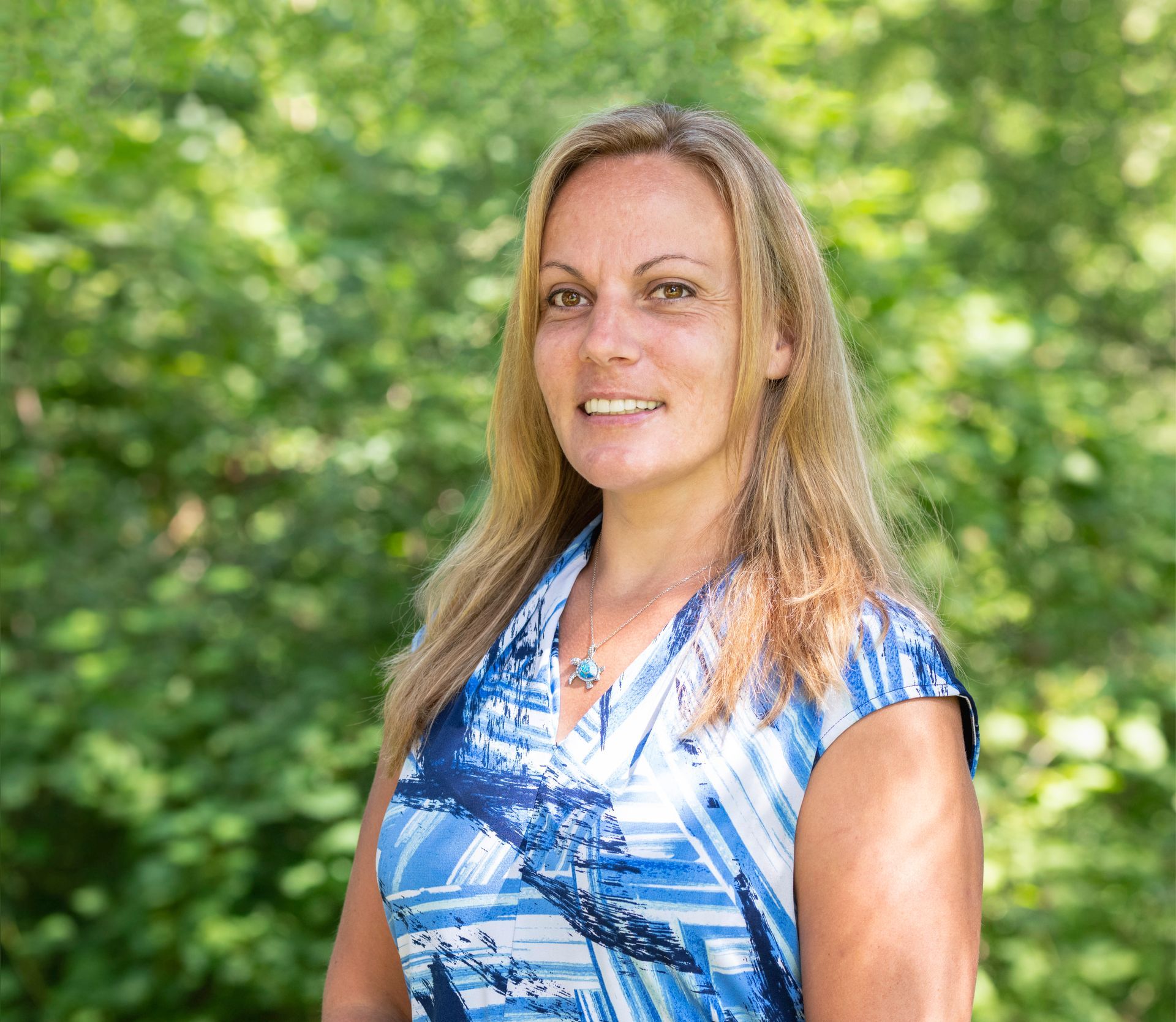 Smiling woman in a blue patterned blouse standing outdoors with green foliage behind her Smiling woman in a blue patterned blouse standing outdoors with green foliage behind her
