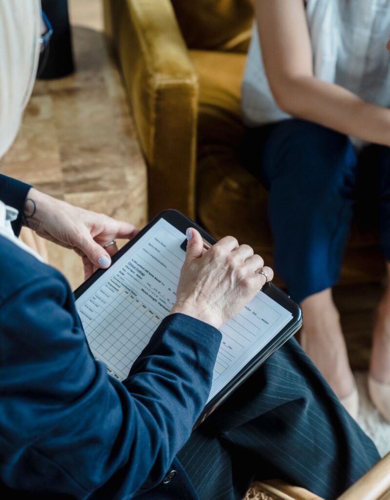 Person taking notes on a tablet at a meeting, with others seated nearby on a sofa.