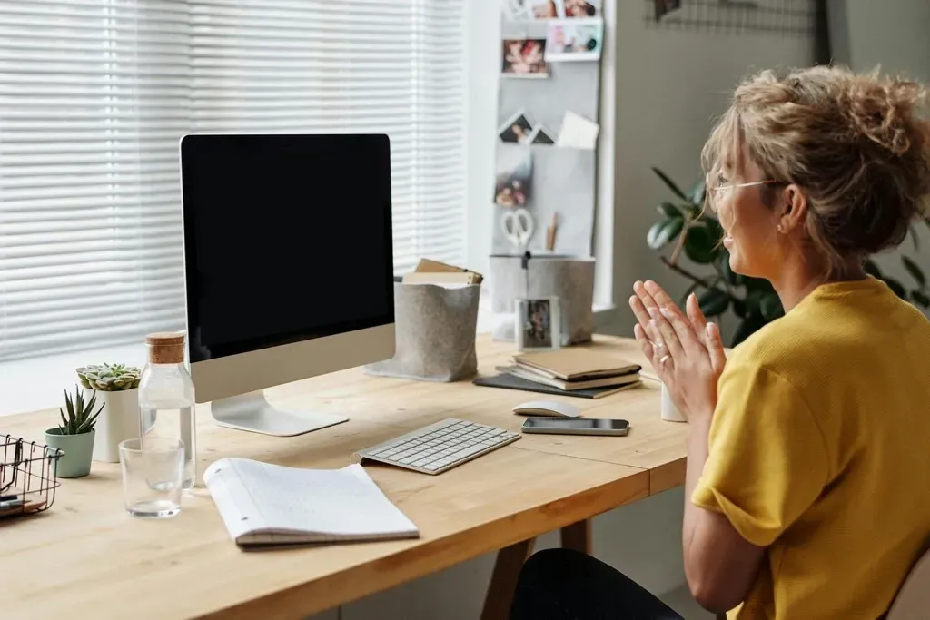 Person seated at a desk, hands clasped, facing a computer in a bright home office