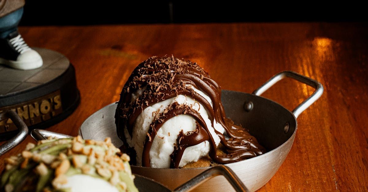 A scoop of vanilla ice cream topped with dark chocolate sauce and shavings, served in a small metal pan on a table. A scoop of vanilla ice cream topped with dark chocolate sauce and shavings, served in a small metal pan on a table.