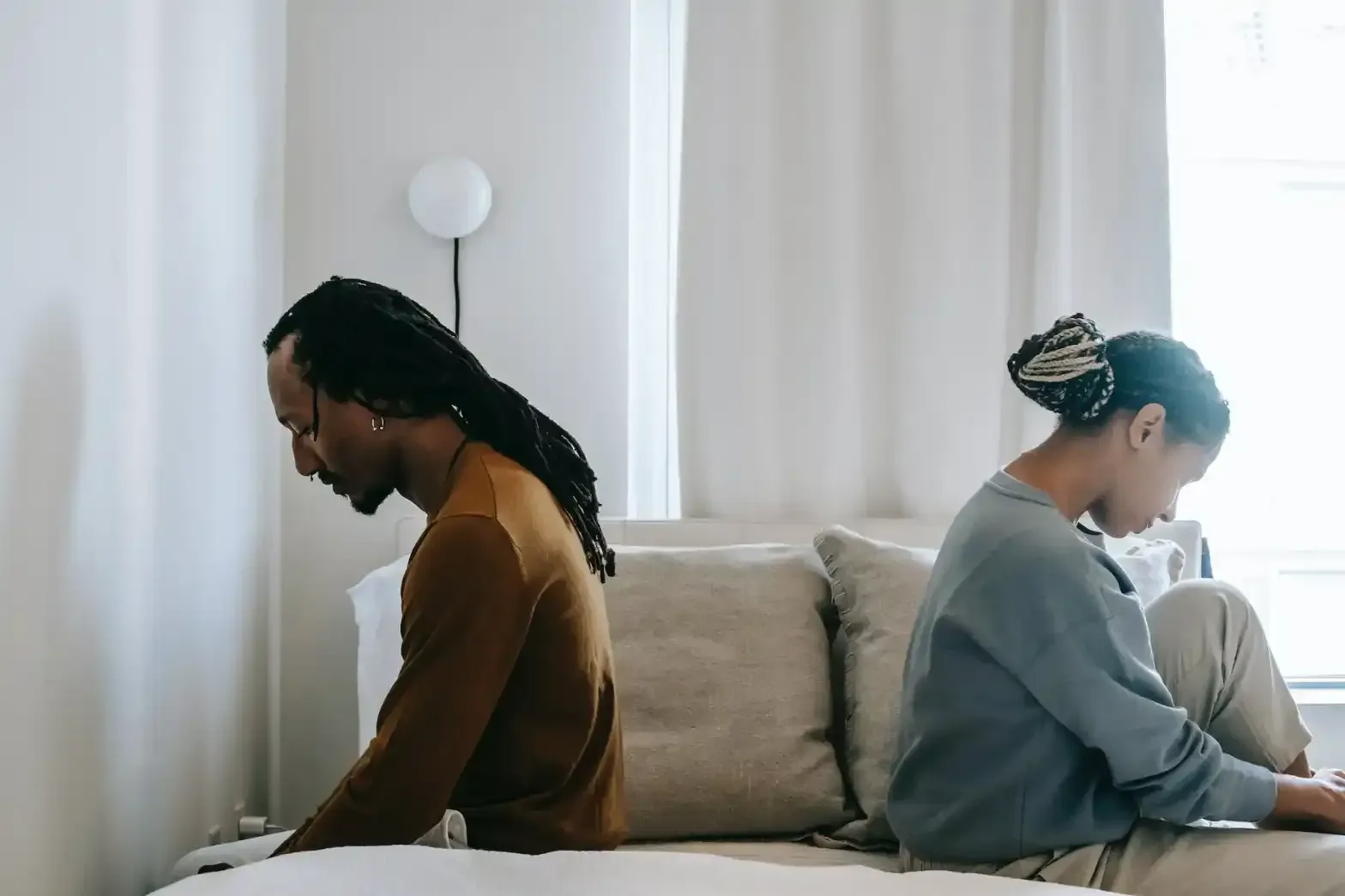 Two people sit back-to-back on a couch in a bright room, each looking down thoughtfully.