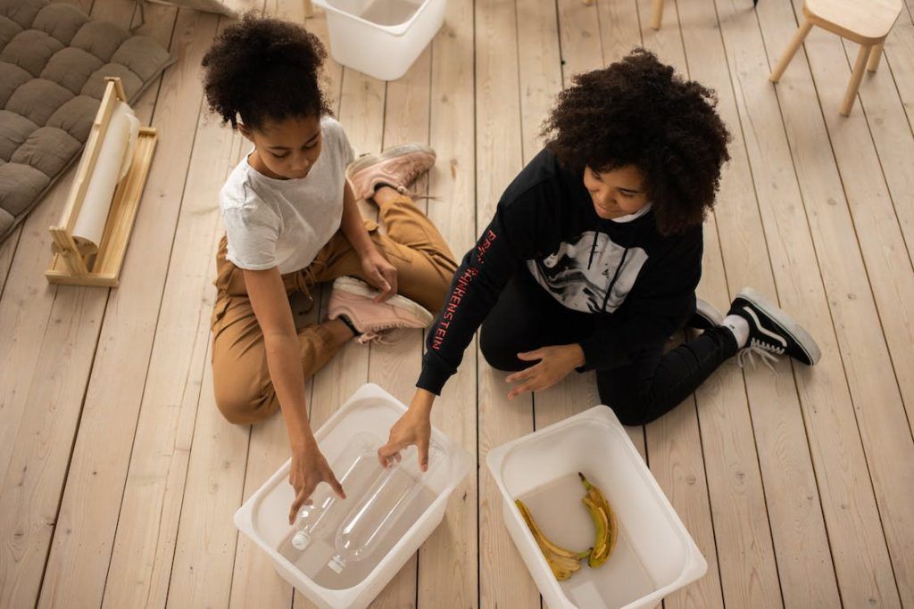 Two people kneel on a wooden floor, arranging items in white bins with a banana nearby.