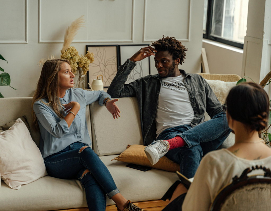 Three people sit in a living room; a woman speaks to a man on a sofa while a third person sits in the foreground. Three people sit in a living room; a woman speaks to a man on a sofa while a third person sits in the foreground.
