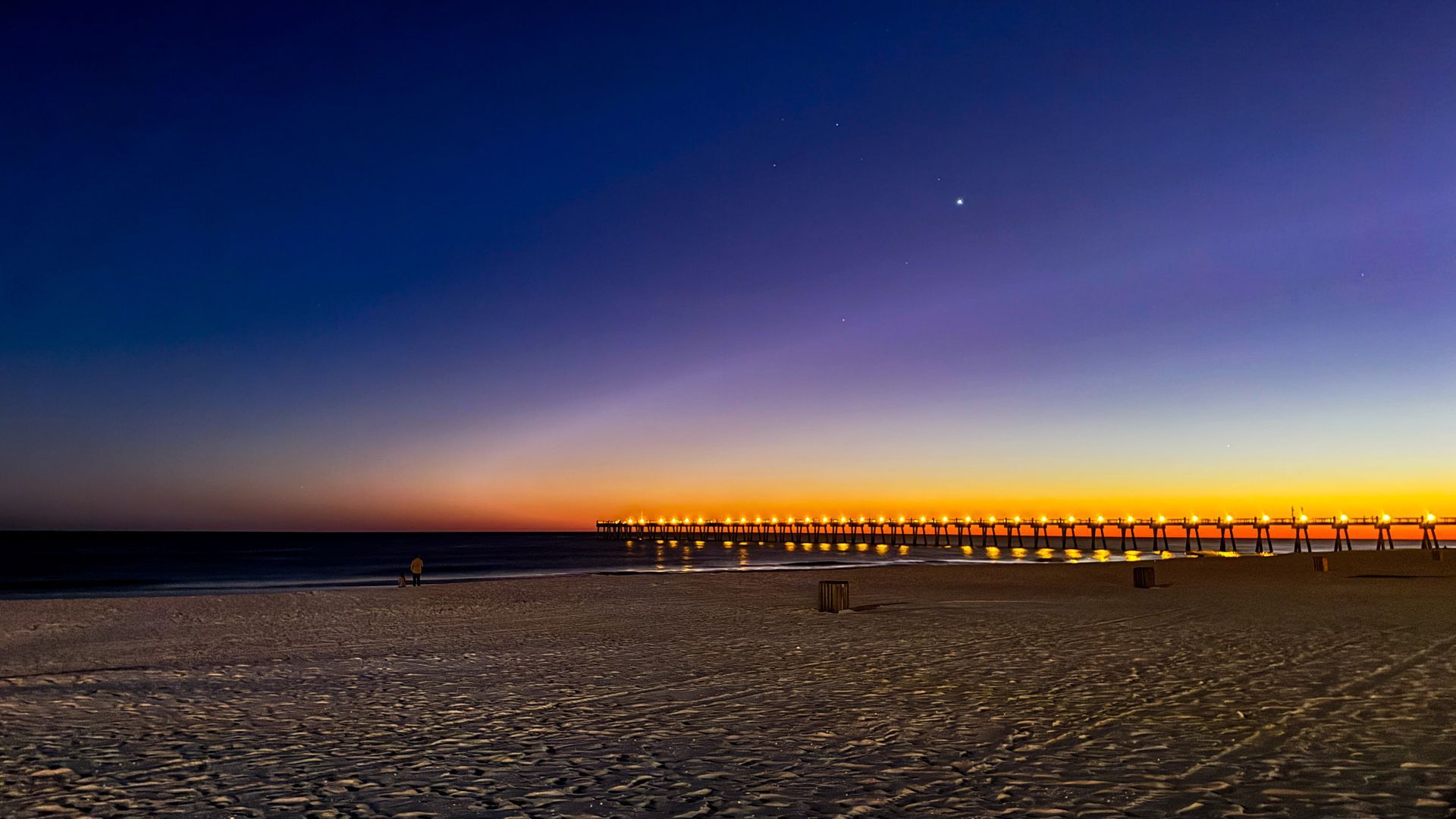Twilight beach with lit pier lights stretching across the horizon under a purple-blue sky
