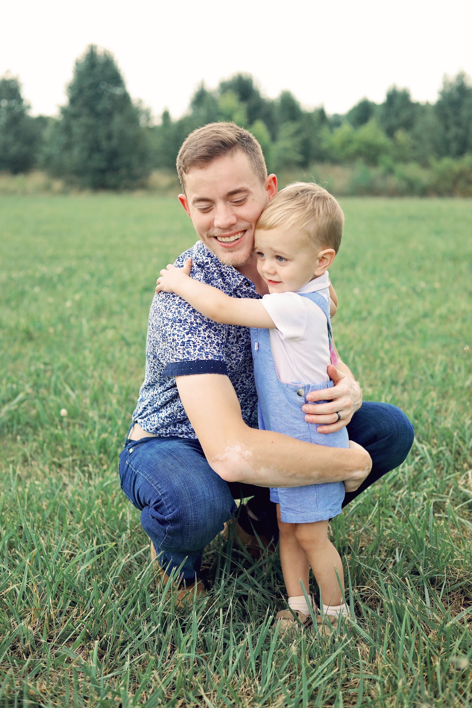Man crouching in a grassy field, smiling while hugging a small child outdoors.