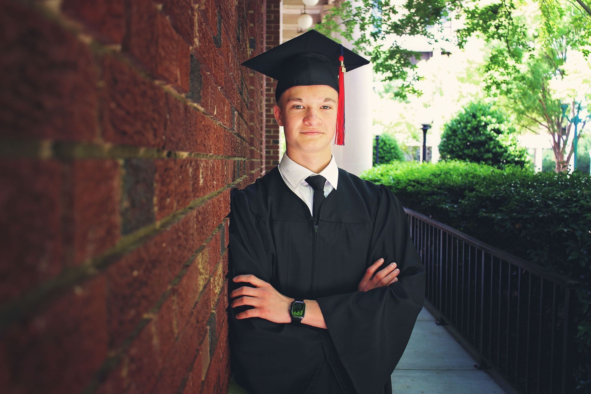 Graduate in black cap and gown leaning against a brick wall outdoors, arms crossed.