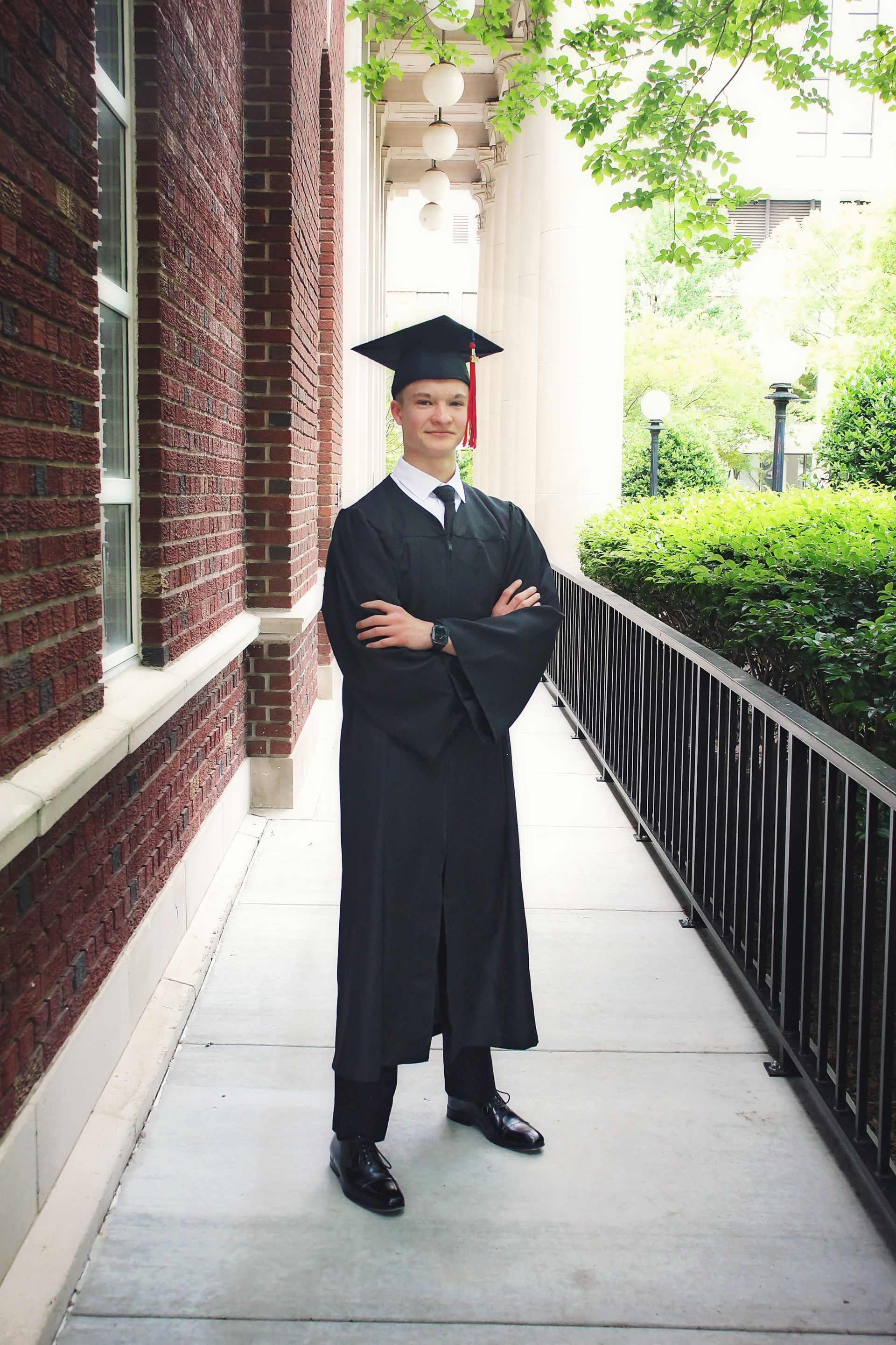 Graduate in black cap and gown standing on a sidewalk beside a brick building and black railing.