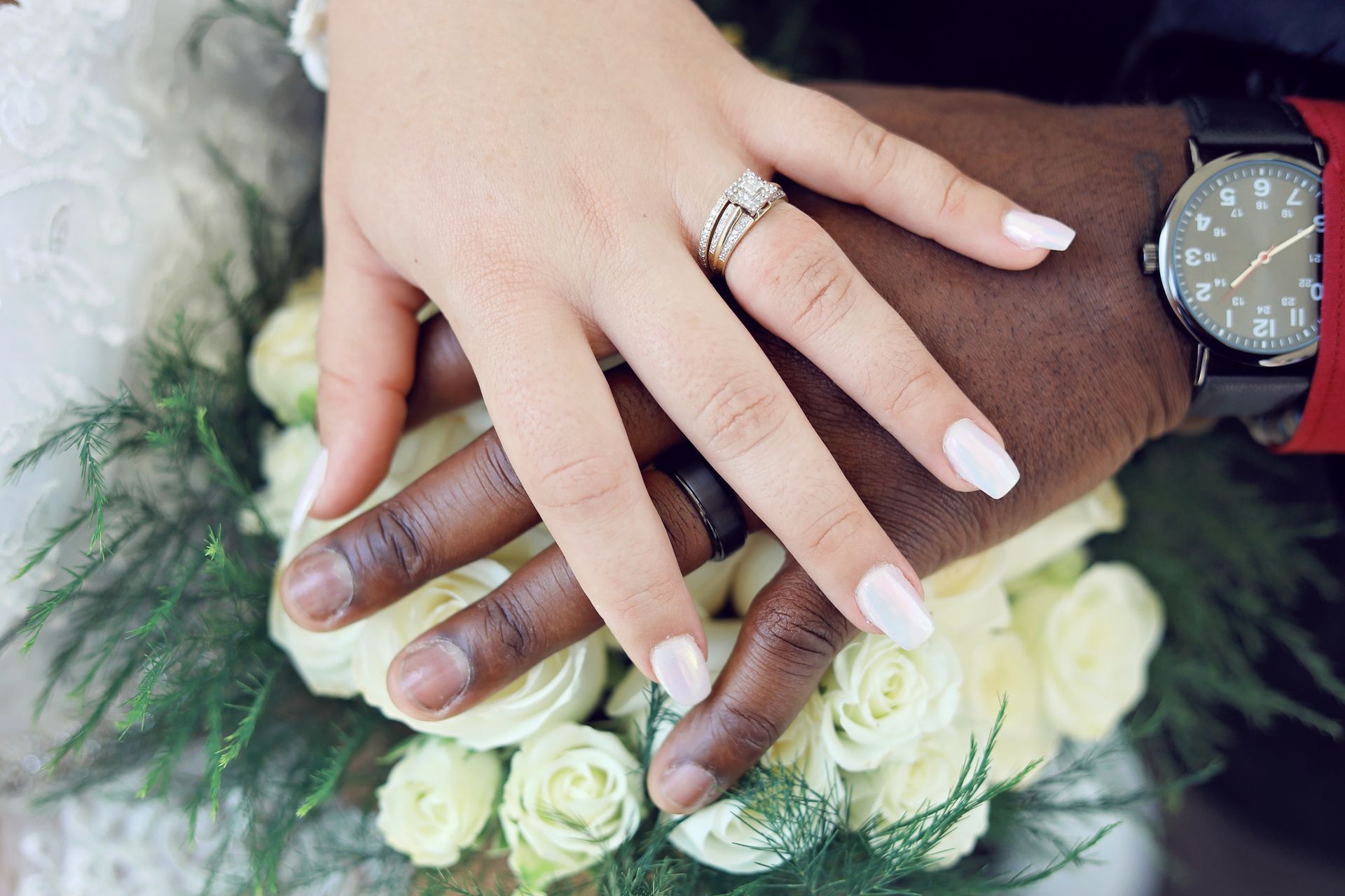 Hands with wedding rings resting on a bouquet of white roses, with a watch visible at the wrist.