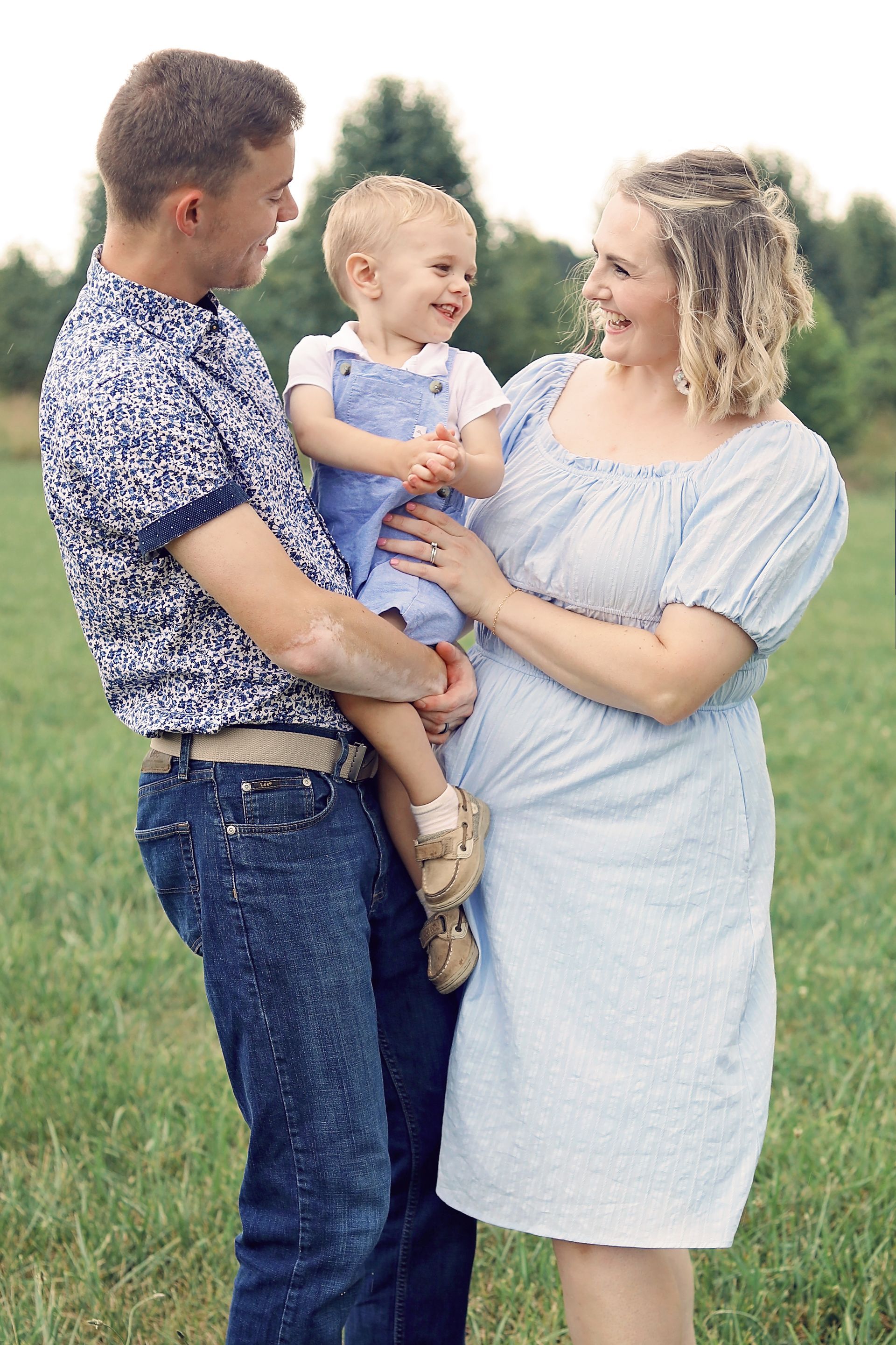 A couple holding a smiling child in a grassy field, dressed in casual summer clothes.