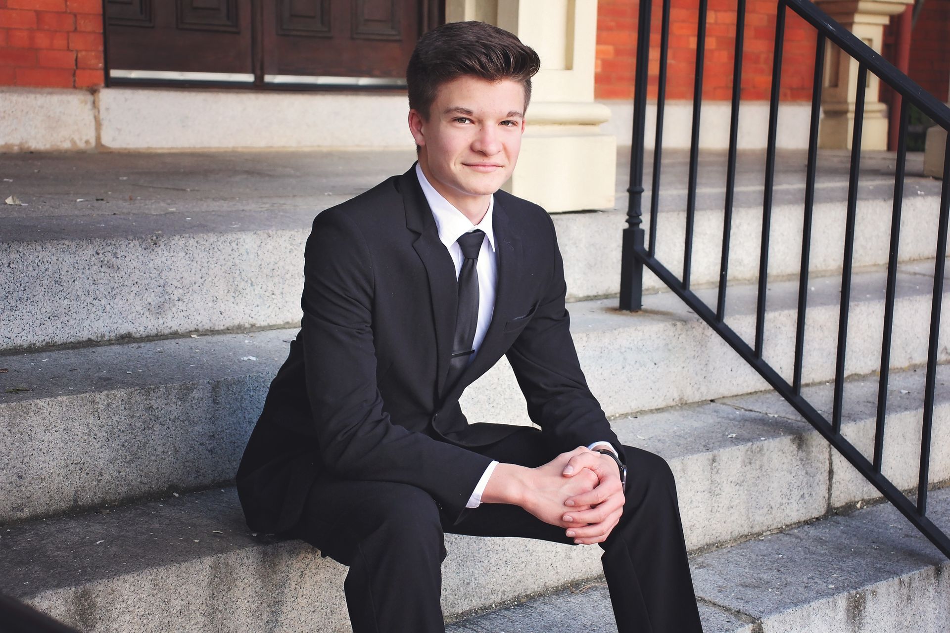 Man in a black suit sitting on stone steps, smiling beside a stair railing.