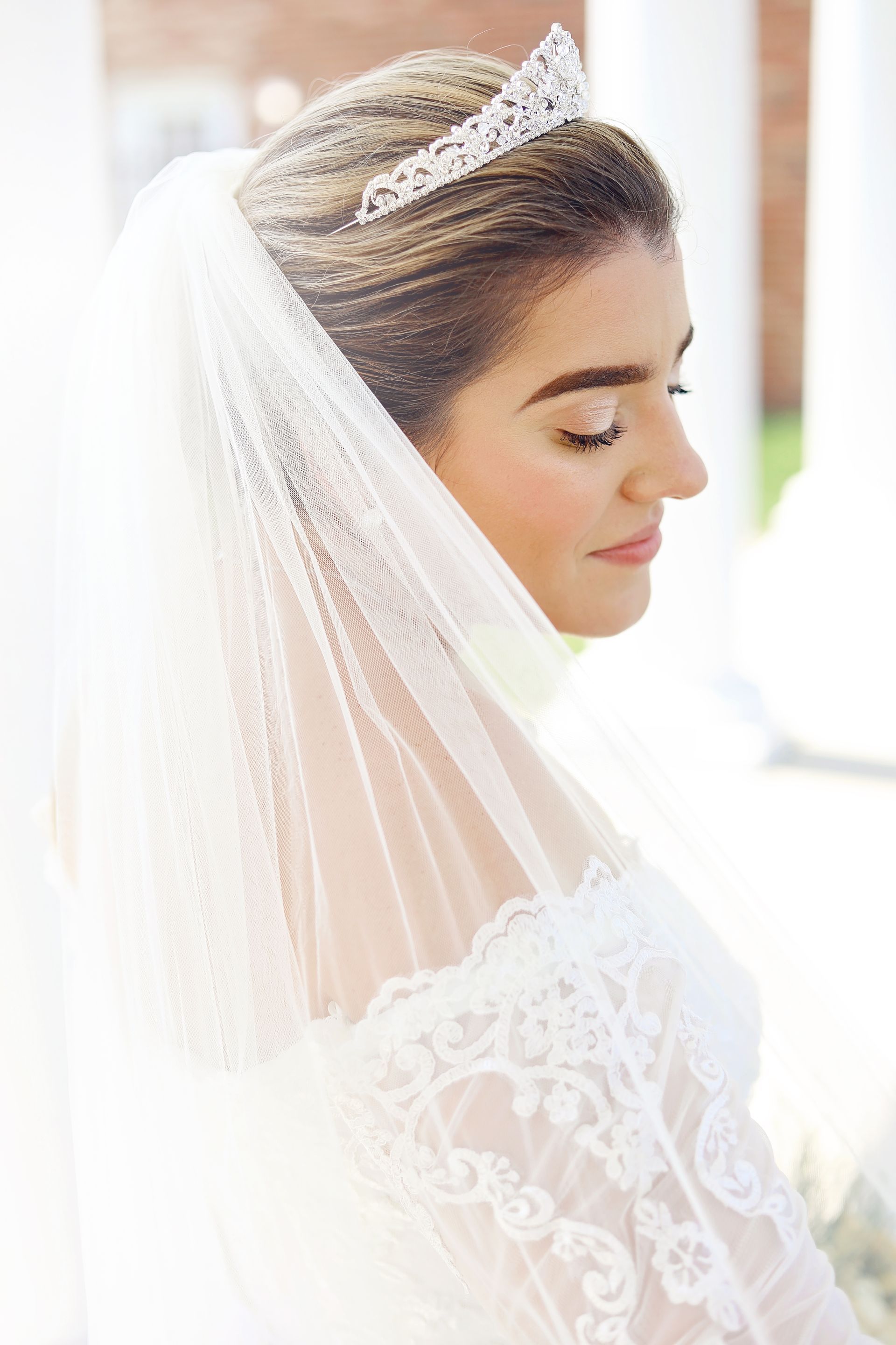 Bride in white veil and tiara, shown in profile with eyes closed, against a bright background