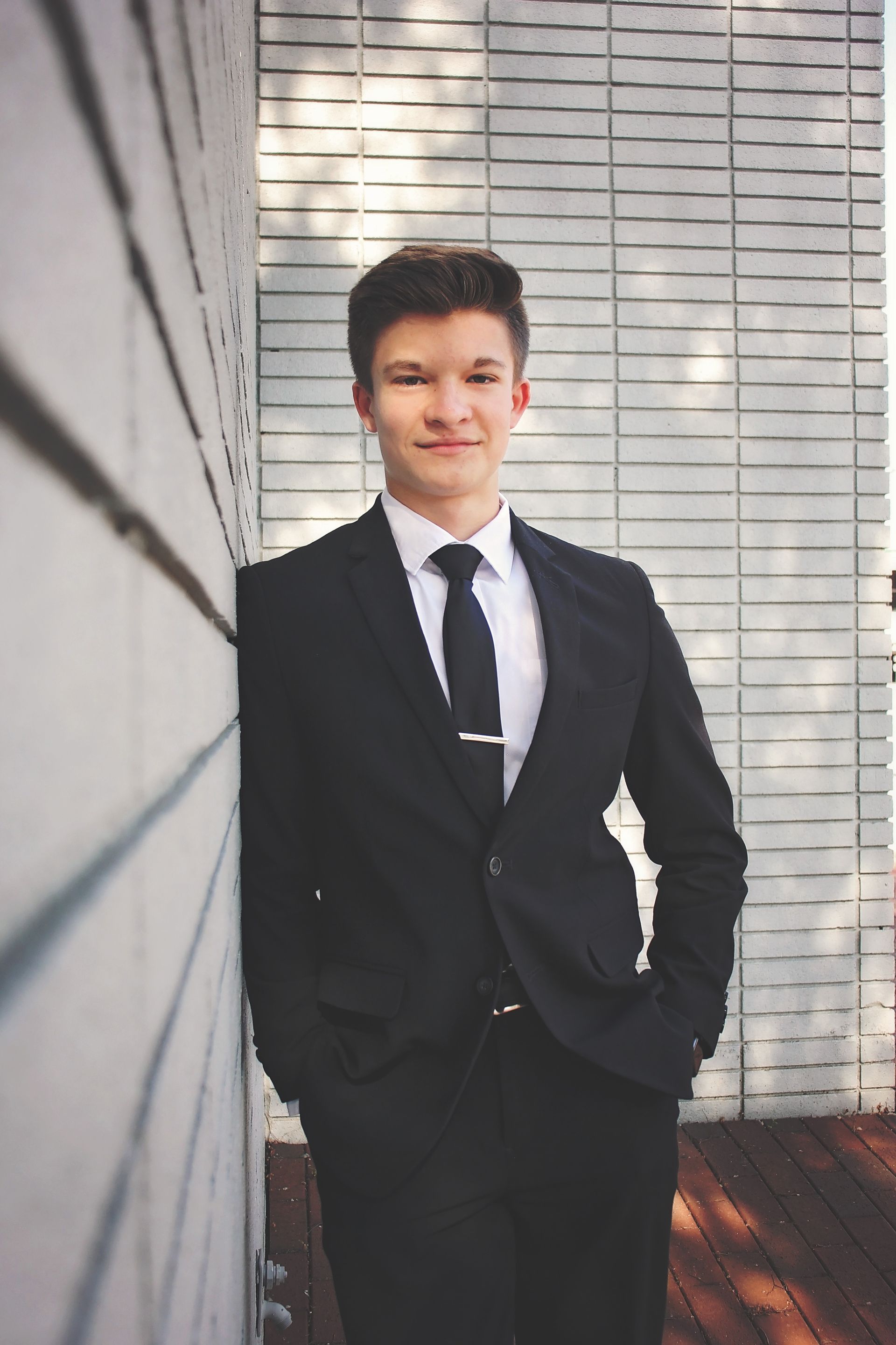 Man in a black suit standing by a brick wall, hands in pockets, with a slight smile.
