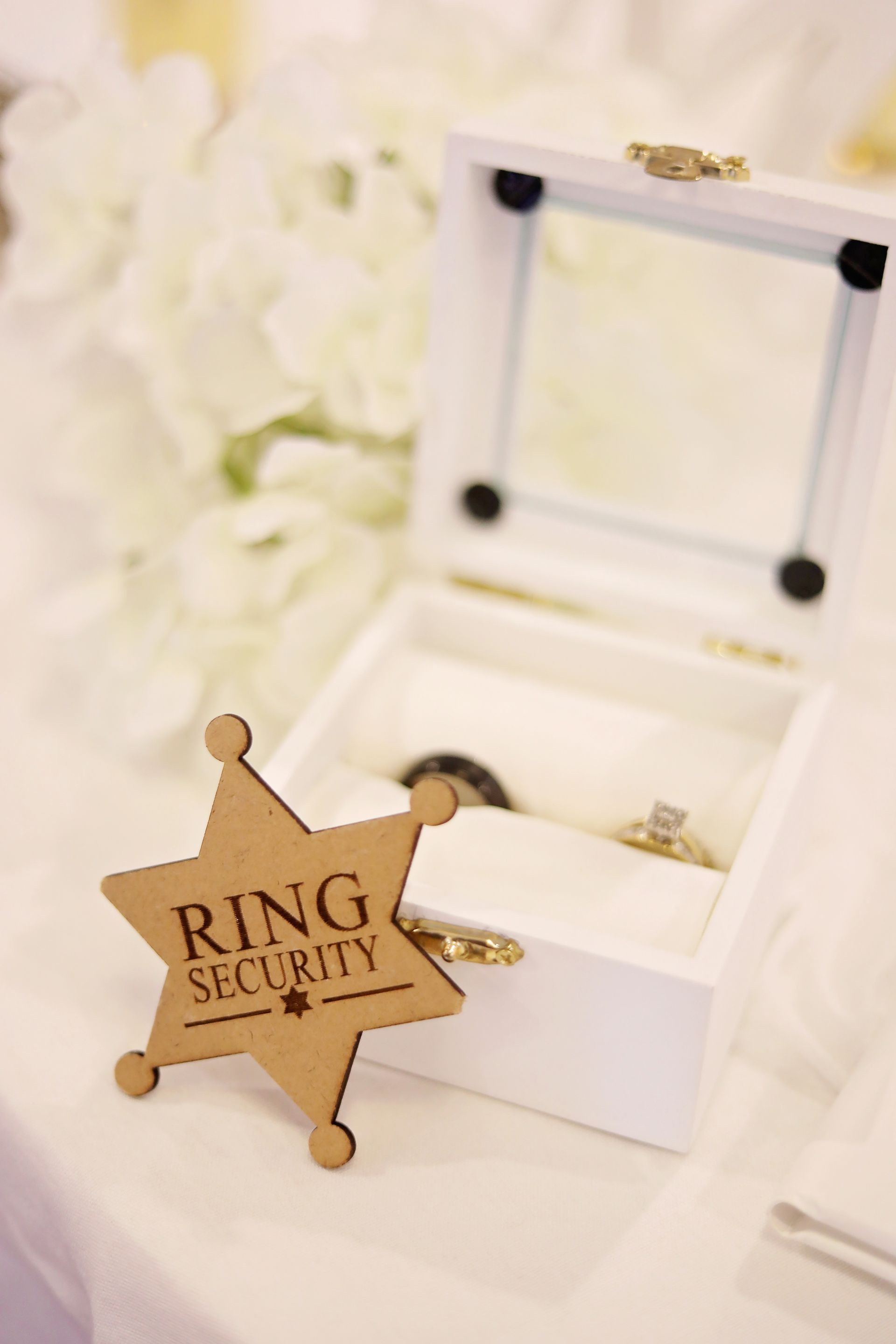 Gold ring in white jewelry box beside a star-shaped “Ring Security” sign and white flowers