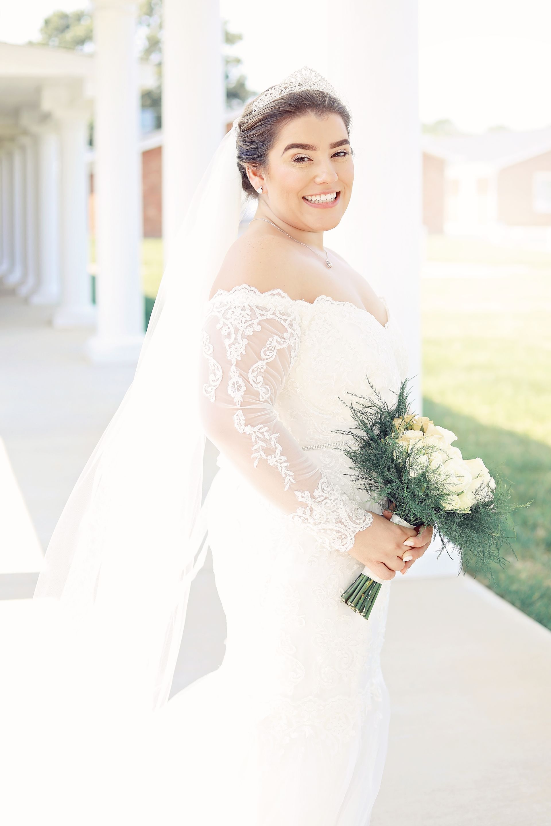 Bride in a white lace wedding gown holding a bouquet outdoors, smiling over her shoulder.