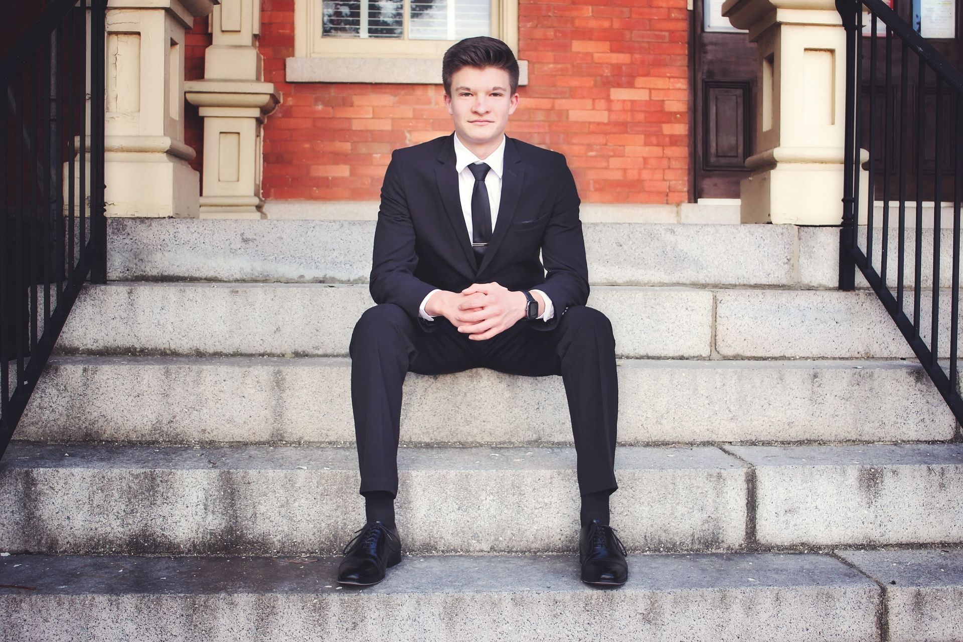 Man in a black suit sitting on stone steps in front of a brick building