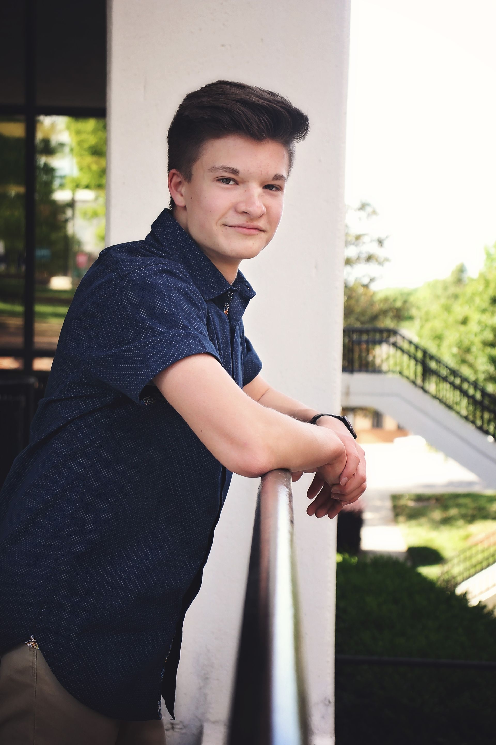 Person leaning on a balcony railing outdoors, wearing a dark blue shirt, with greenery in the background