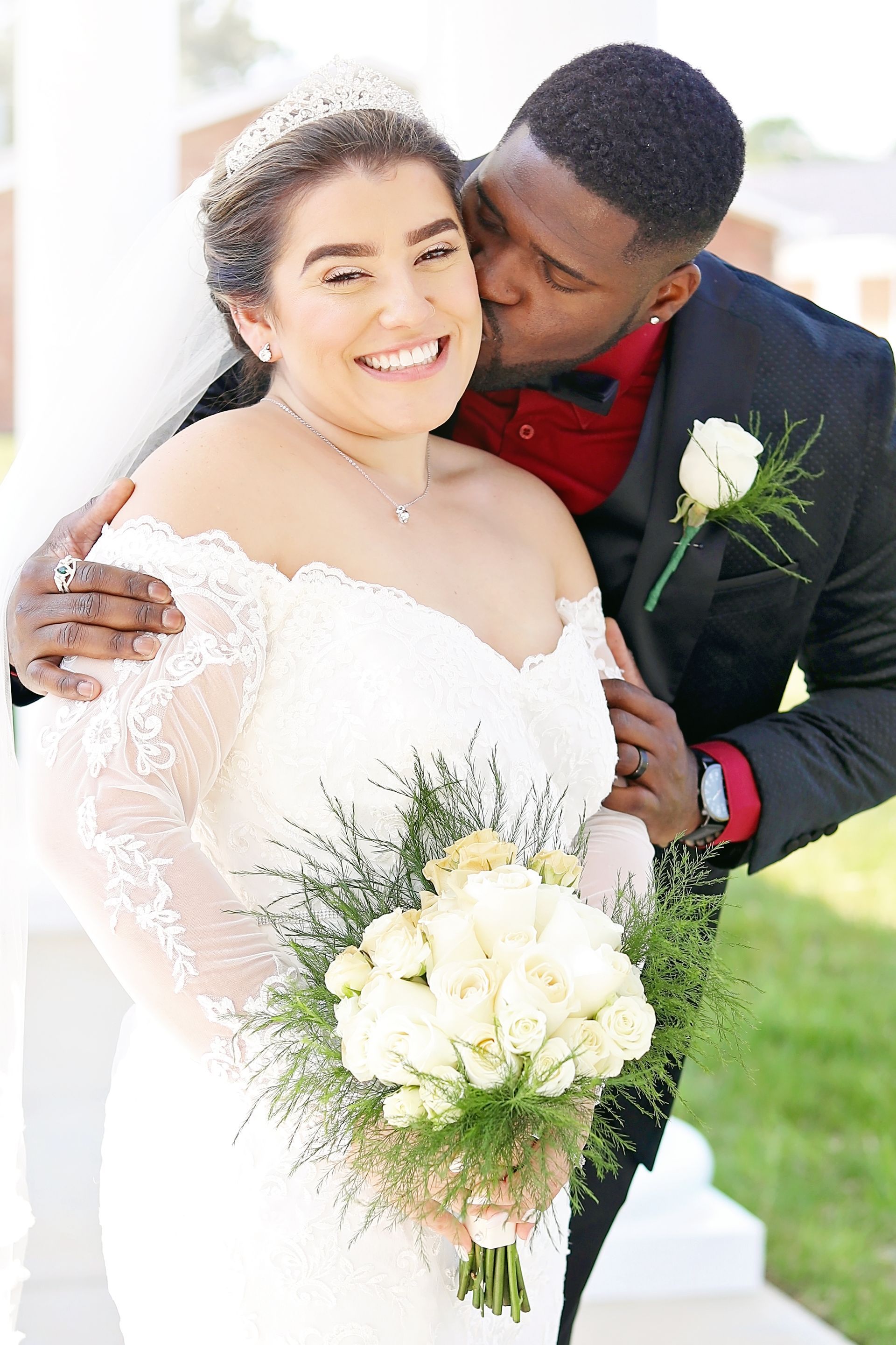 Bride and groom embracing outdoors, smiling with a white bouquet and rose boutonniere.