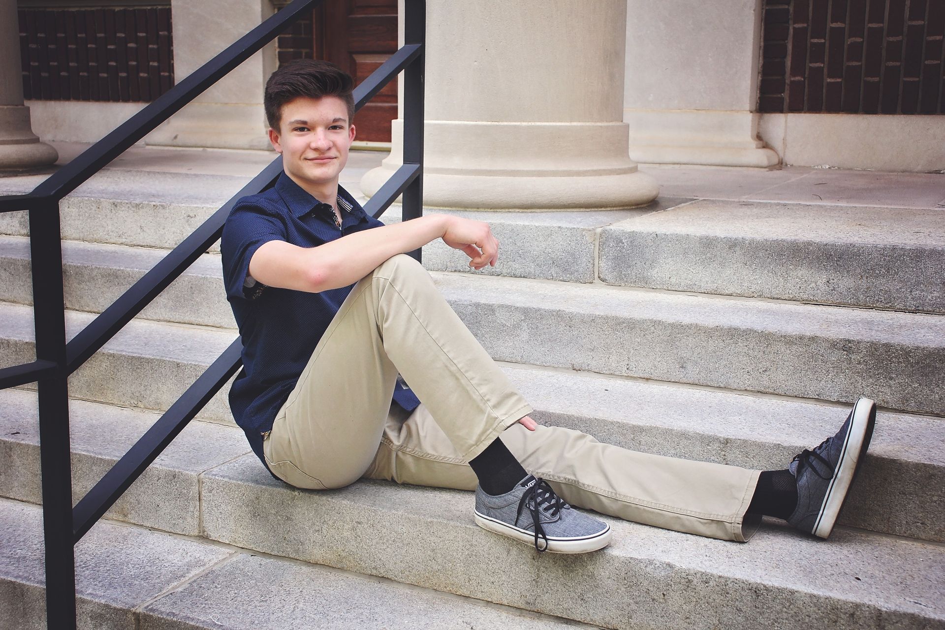 Person sitting on outdoor stone steps, wearing dark shirt and tan pants, smiling at the camera.