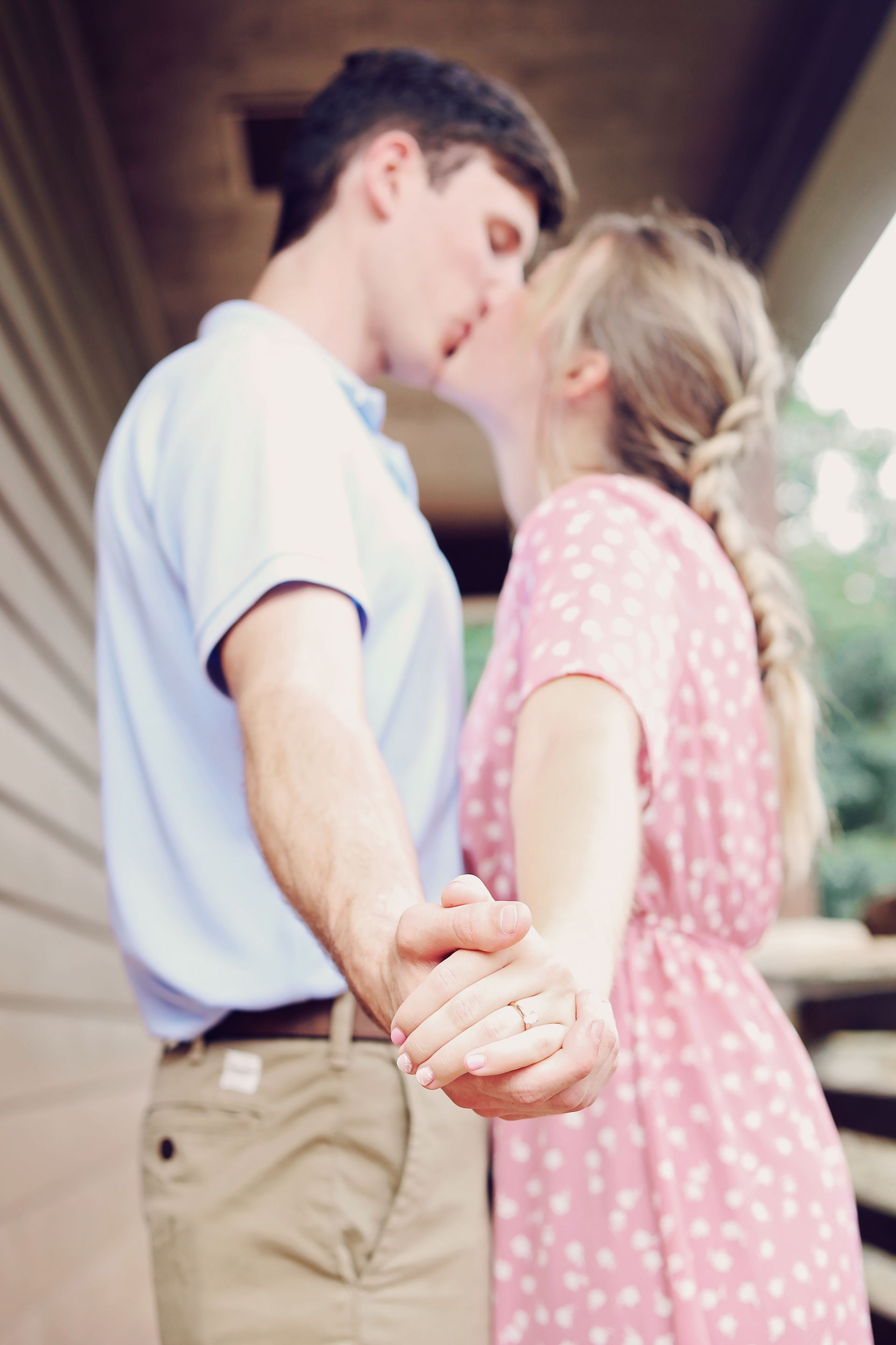 Couple kissing while holding hands on a porch, with the woman in a pink dress and the man in a white shirt.