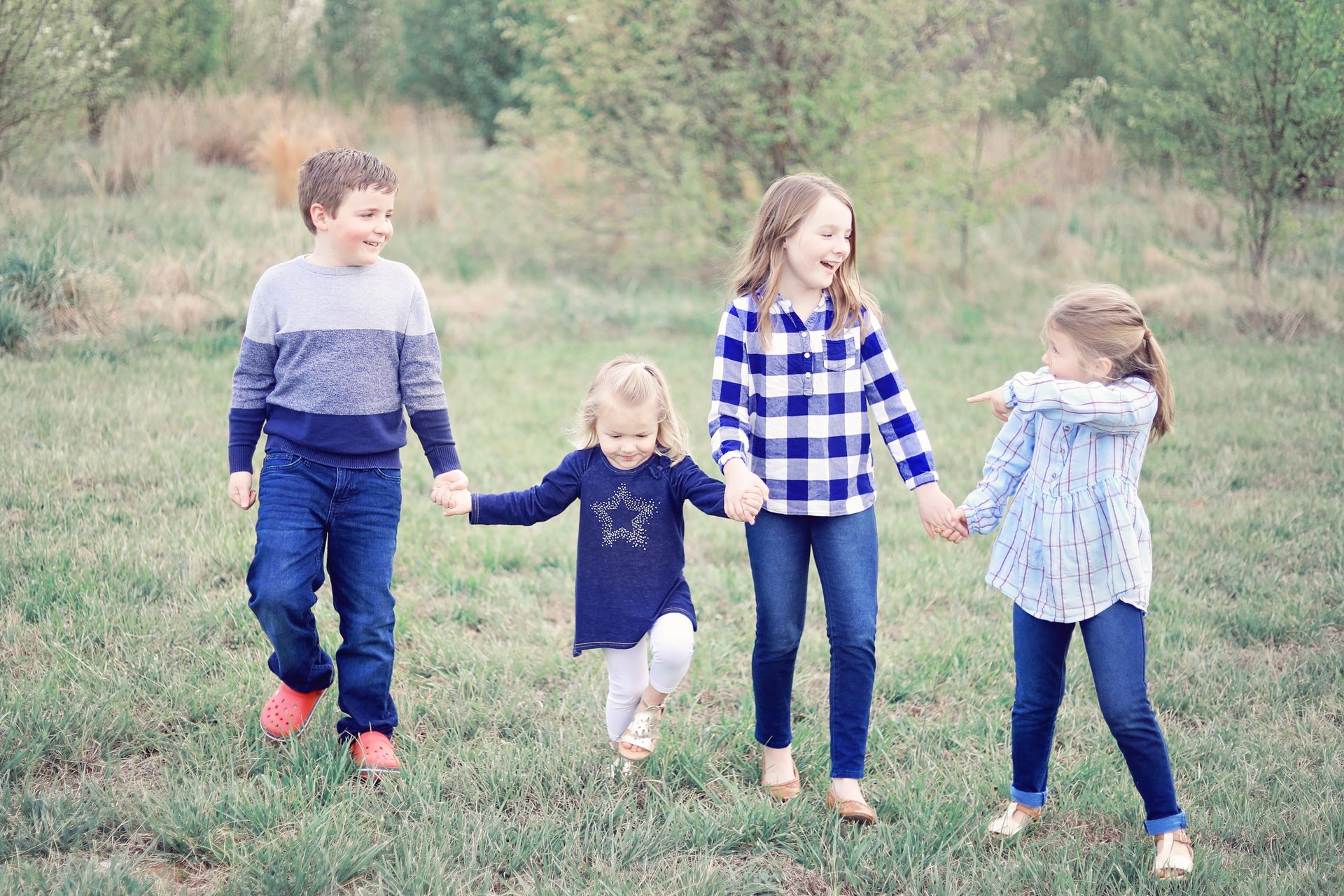 Four children holding hands and walking through a grassy field, wearing blue and white clothes