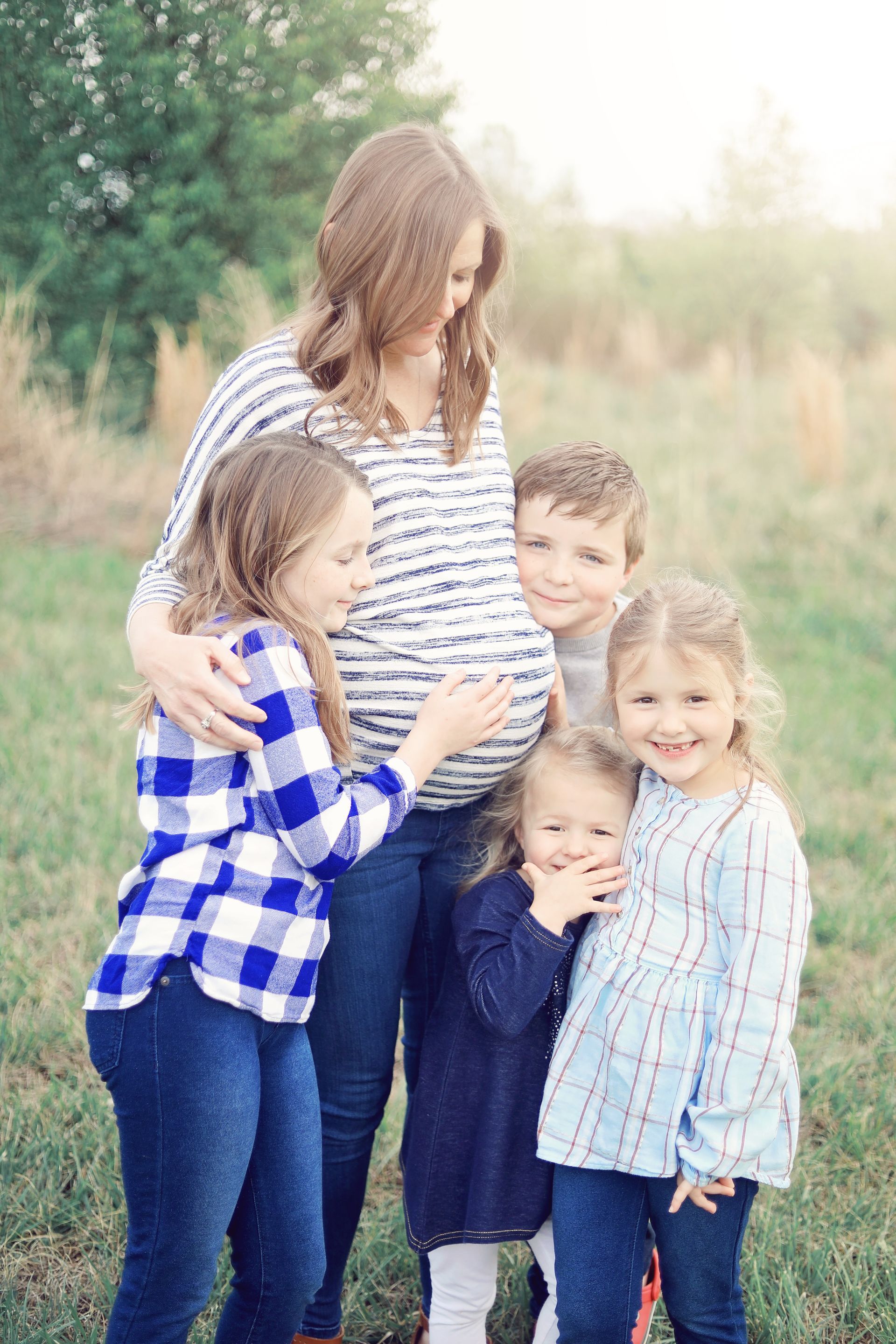 Woman and four children standing together outdoors in a grassy field, smiling and hugging.