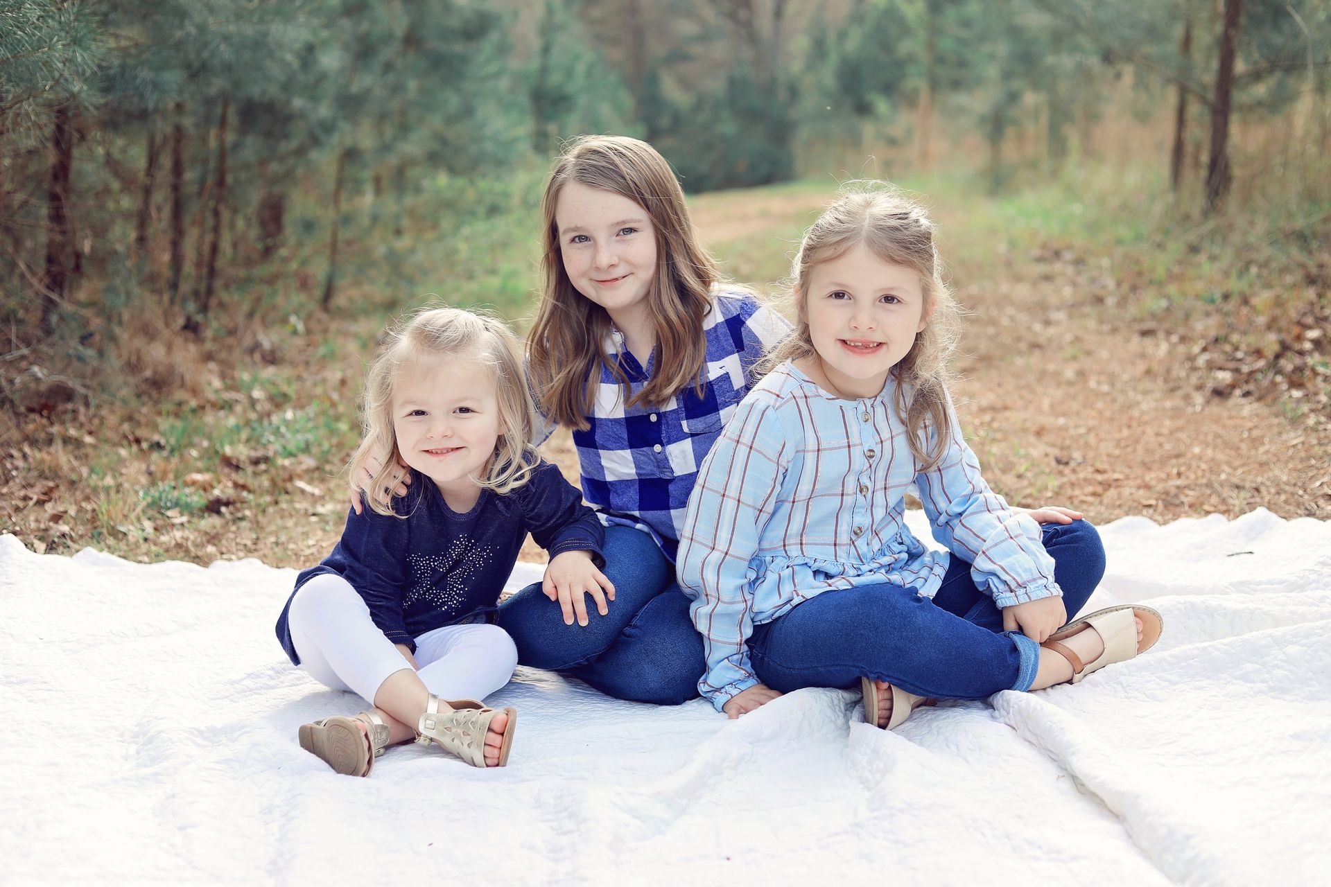 Three children sitting on a white blanket in a wooded area, smiling at the camera.