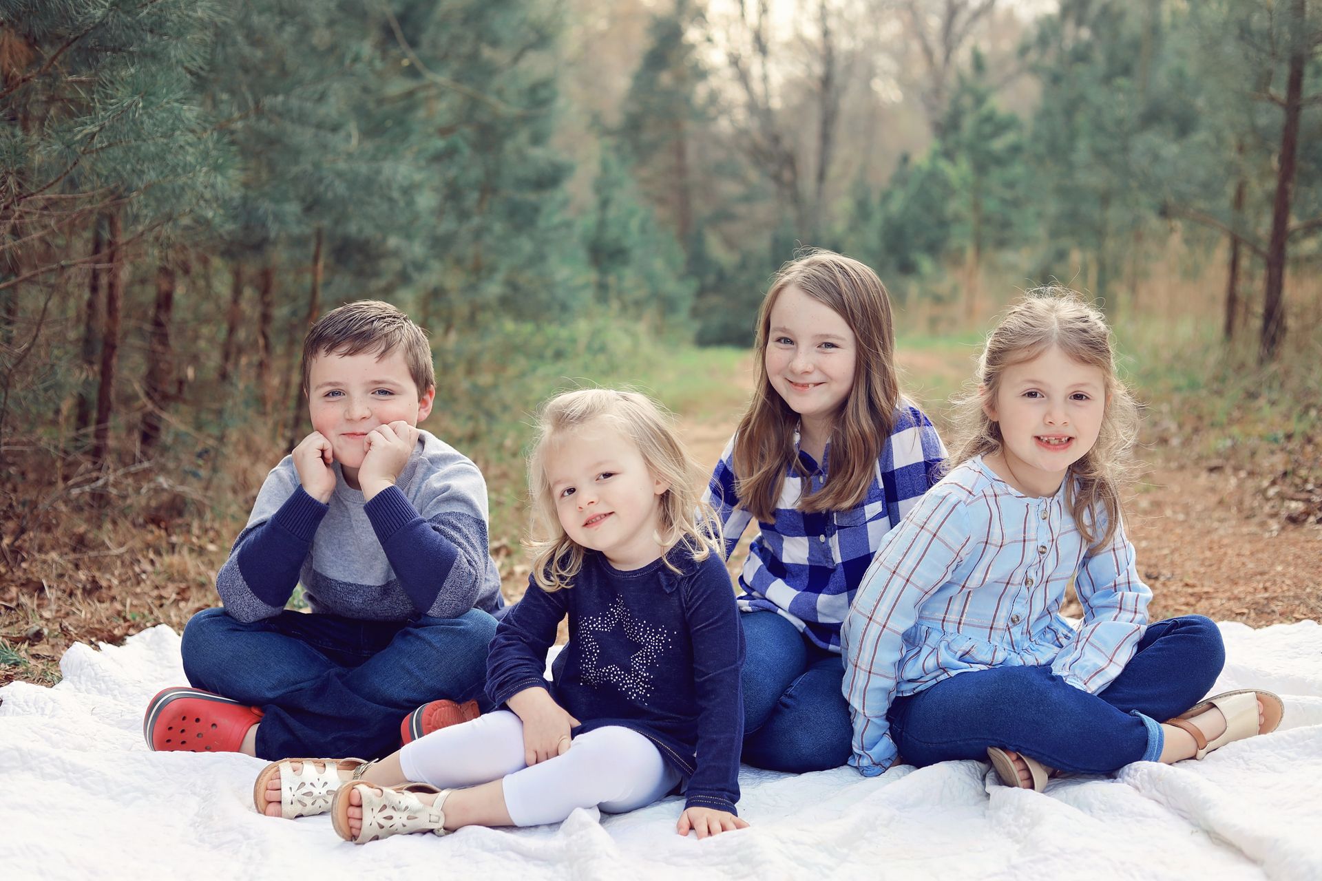 Four children sitting on a white blanket in a wooded area, smiling at the camera.