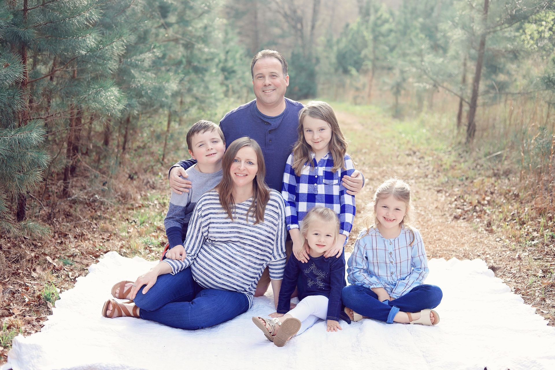 Family sitting on a white blanket outdoors in a wooded area, smiling at the camera.