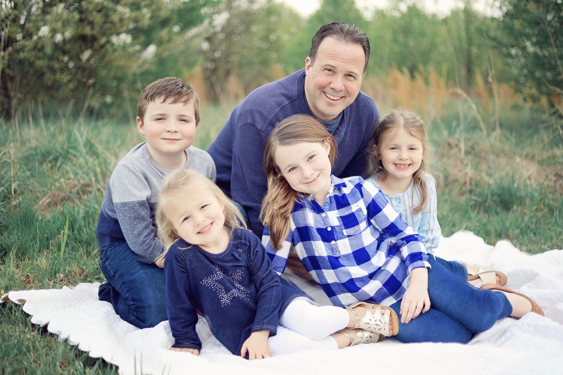 Family sitting together on a white blanket outdoors, smiling at the camera.