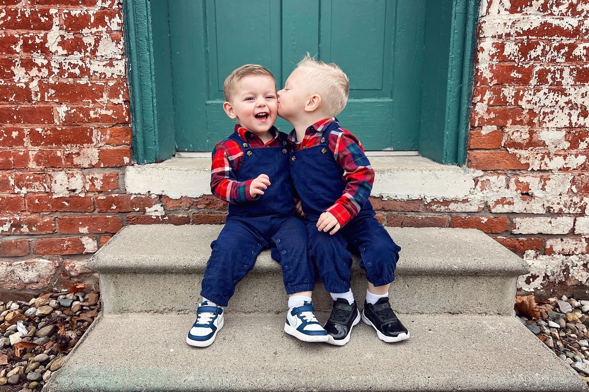 Two smiling toddlers in blue overalls sit on steps in front of a red brick wall and teal door.