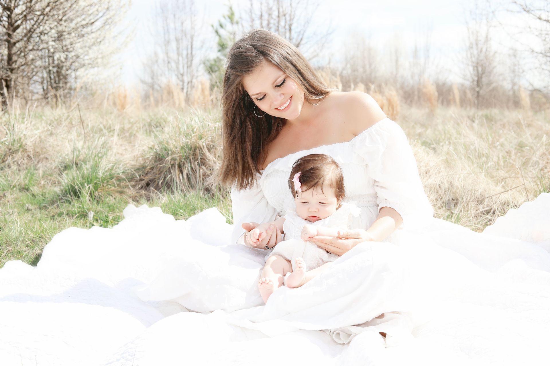 Woman holding a baby on a white blanket in a sunlit field, both dressed in white.