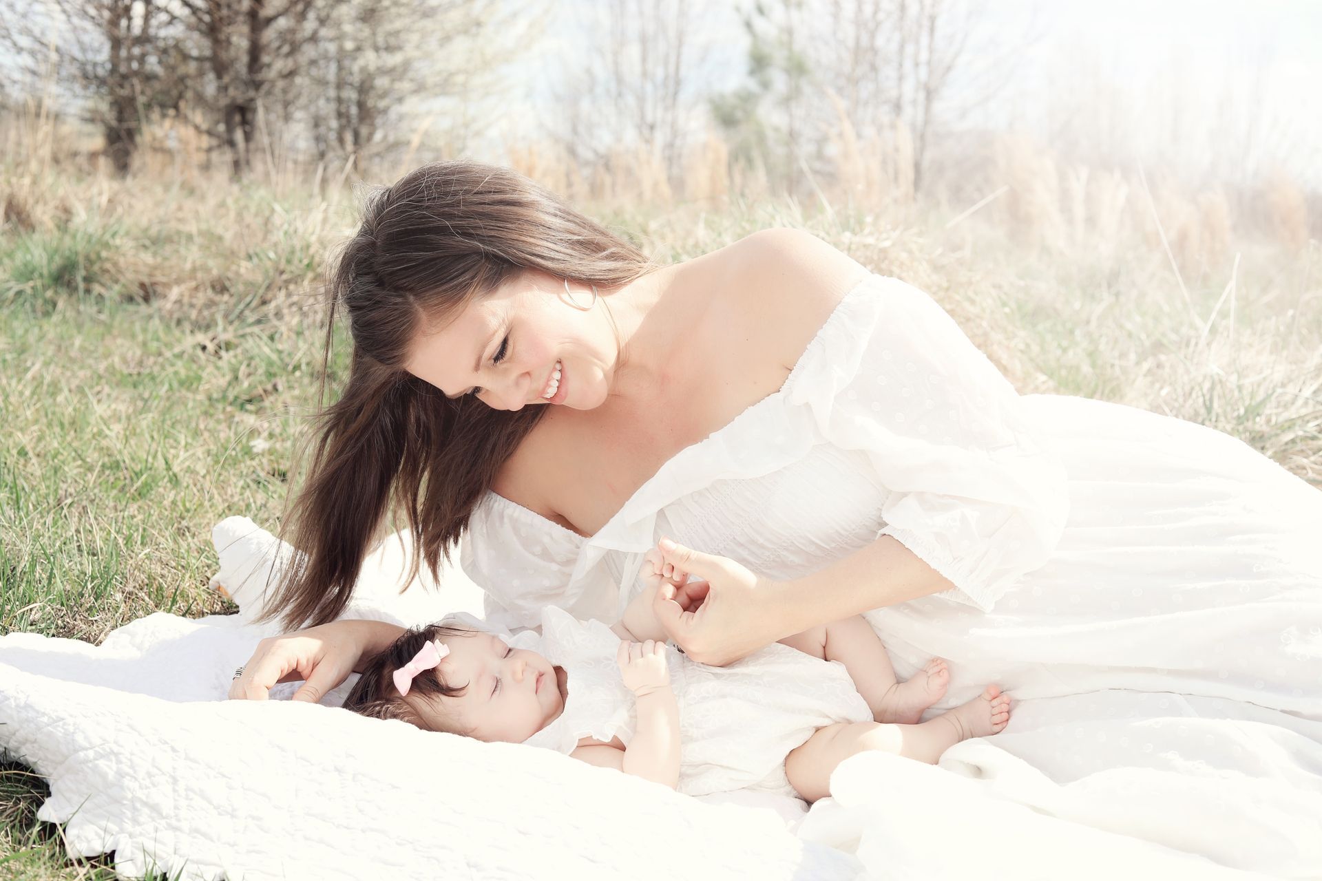 Woman in a white dress lying with a baby on a blanket in a bright grassy field
